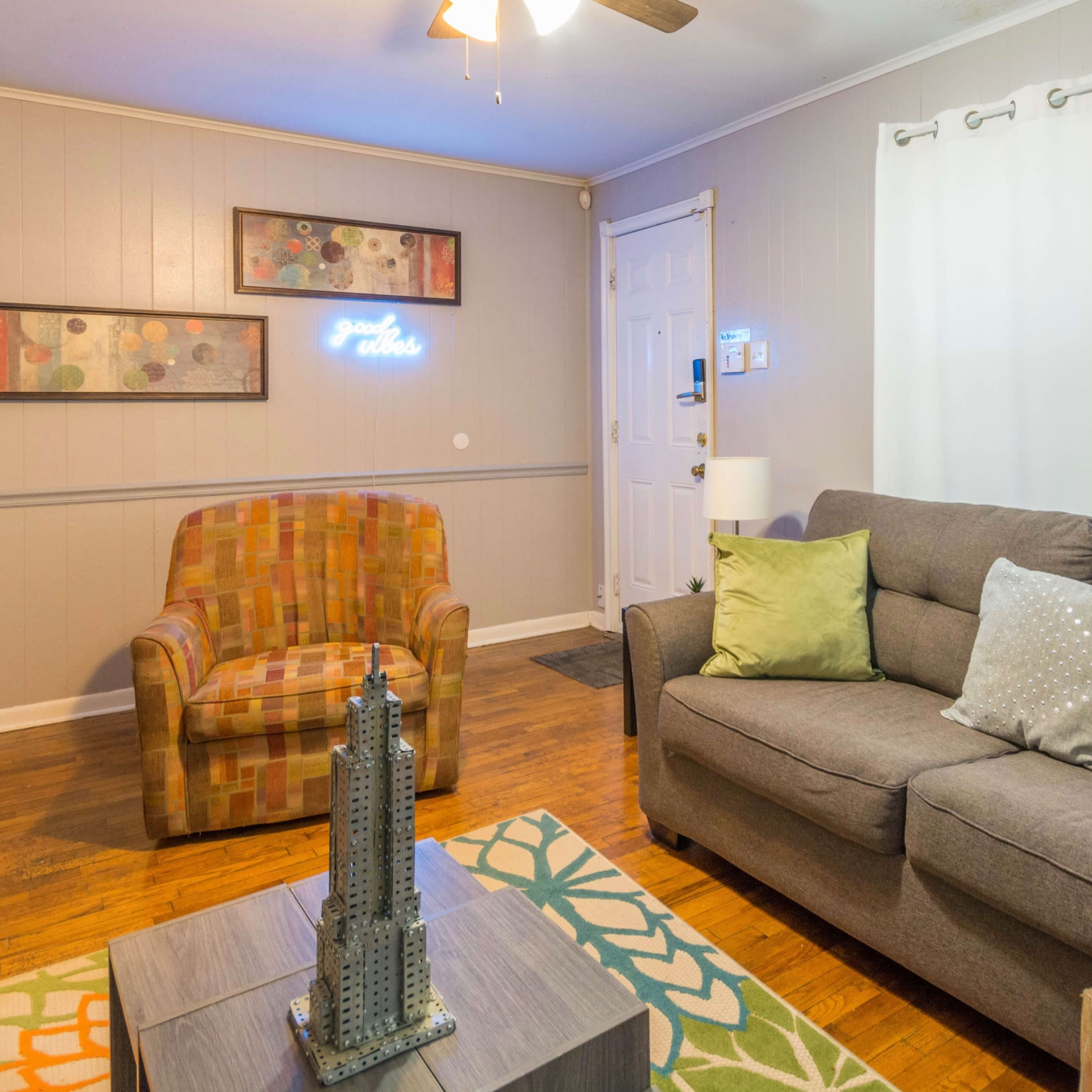 The living room features a gray sofa with green pillows, an accent chair with a checkered pattern, a wooden coffee table, and light-colored walls.