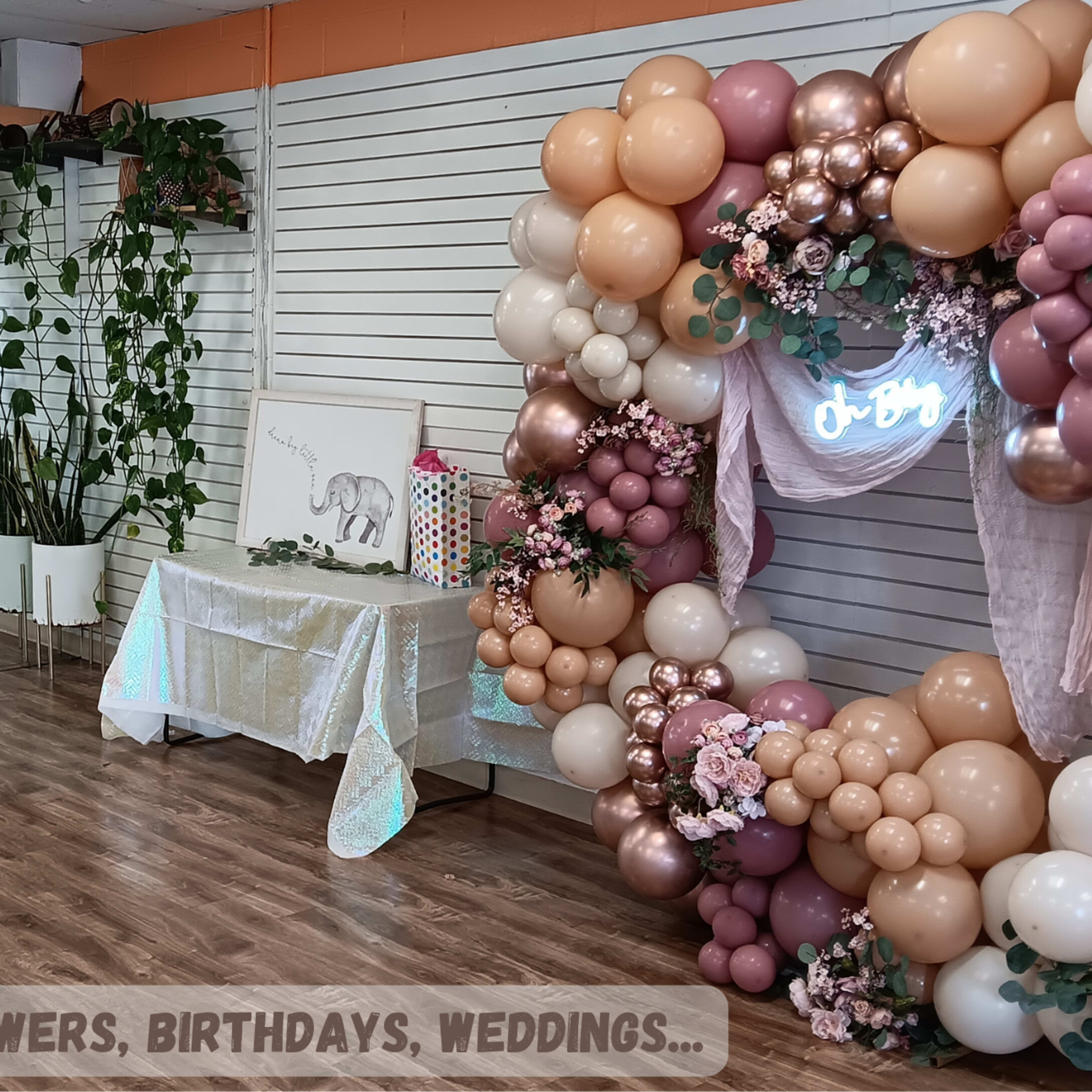 A decorated indoor space featuring tables with desserts and a balloon arch adorned with flowers, designed for events like baby showers and birthdays.