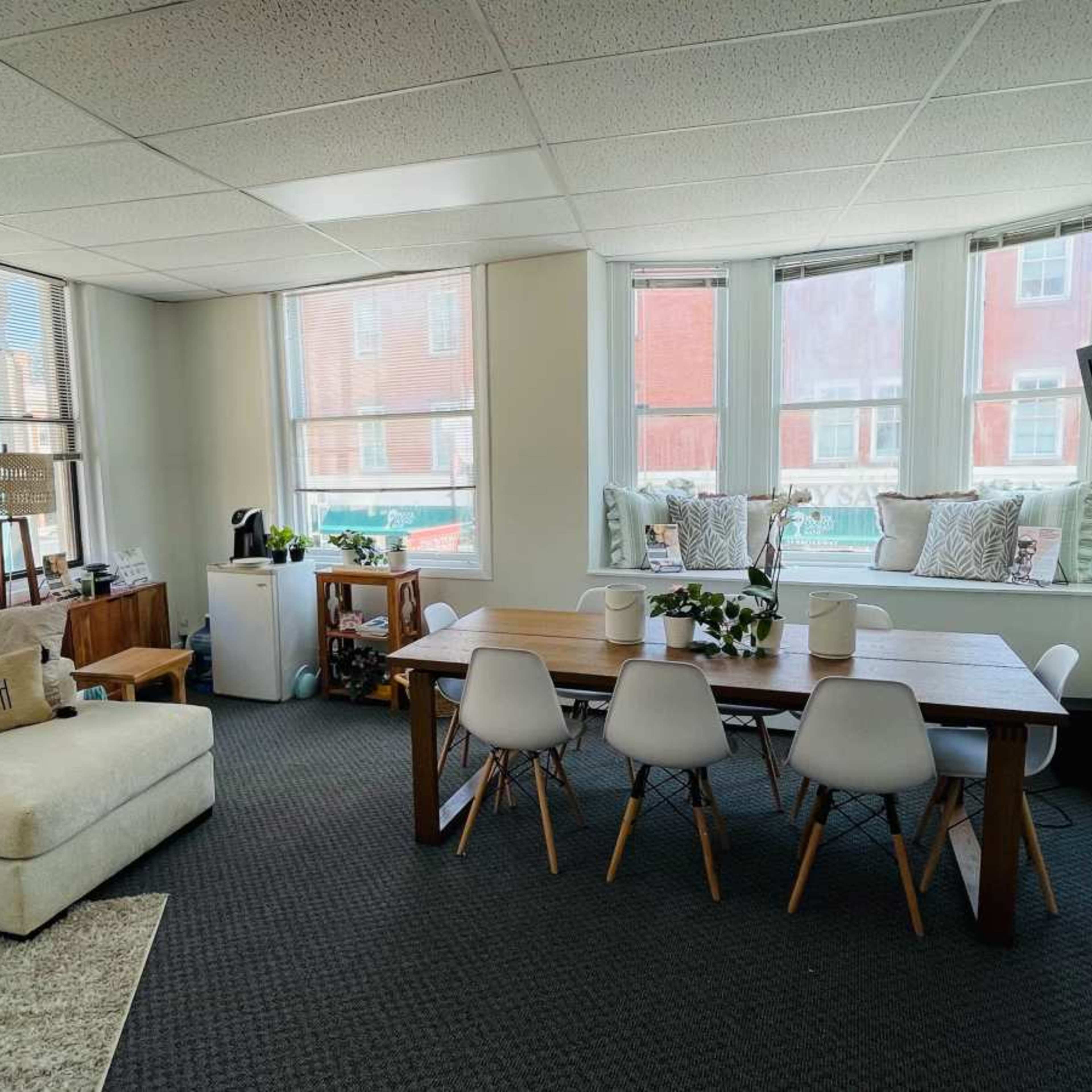 The room features a seating area with a light-colored couch, a wooden dining table surrounded by white chairs, and large windows allowing natural light.