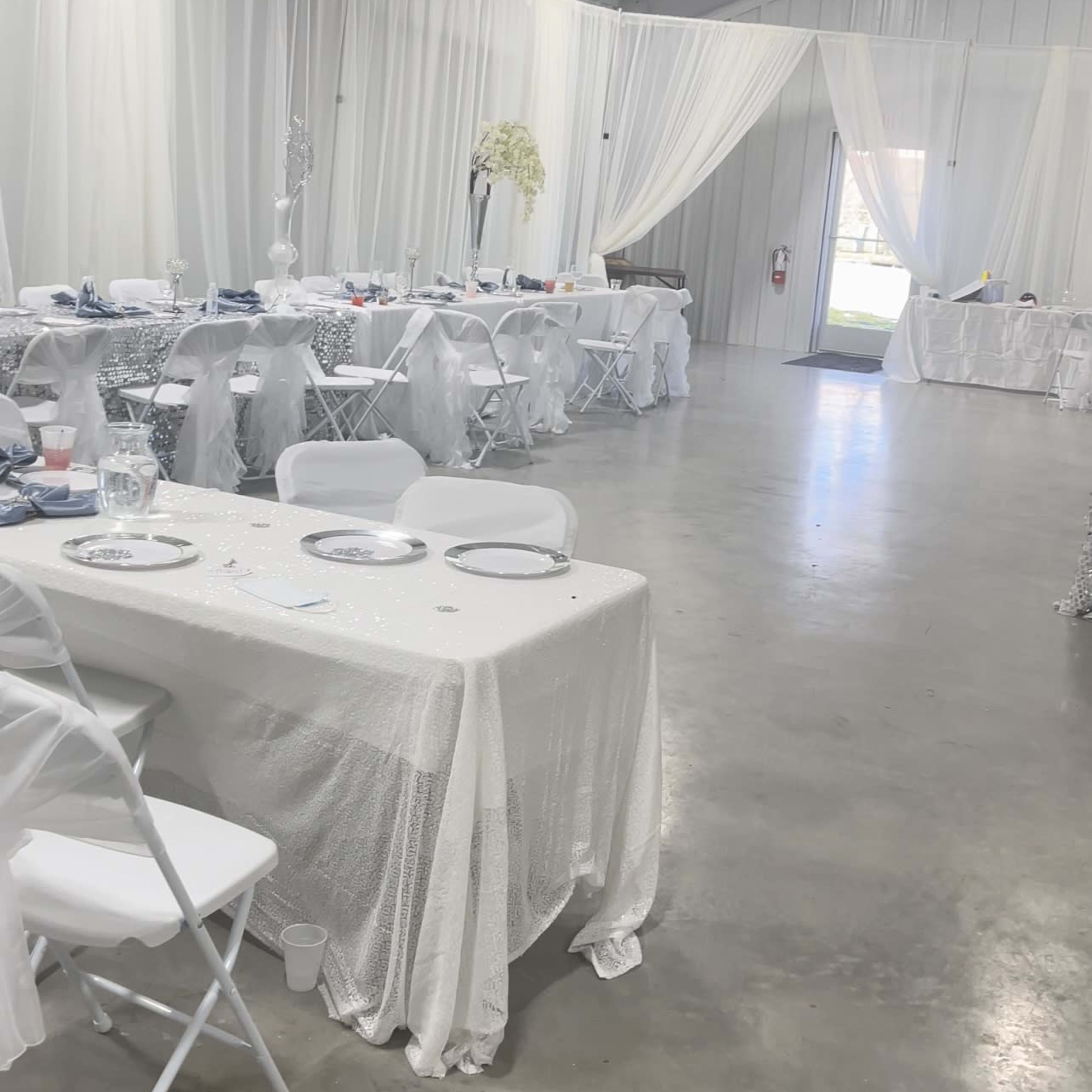 A large indoor event space is set up with white tables and chairs, adorned with decorative tablecloths and surrounded by sheer white drapery.