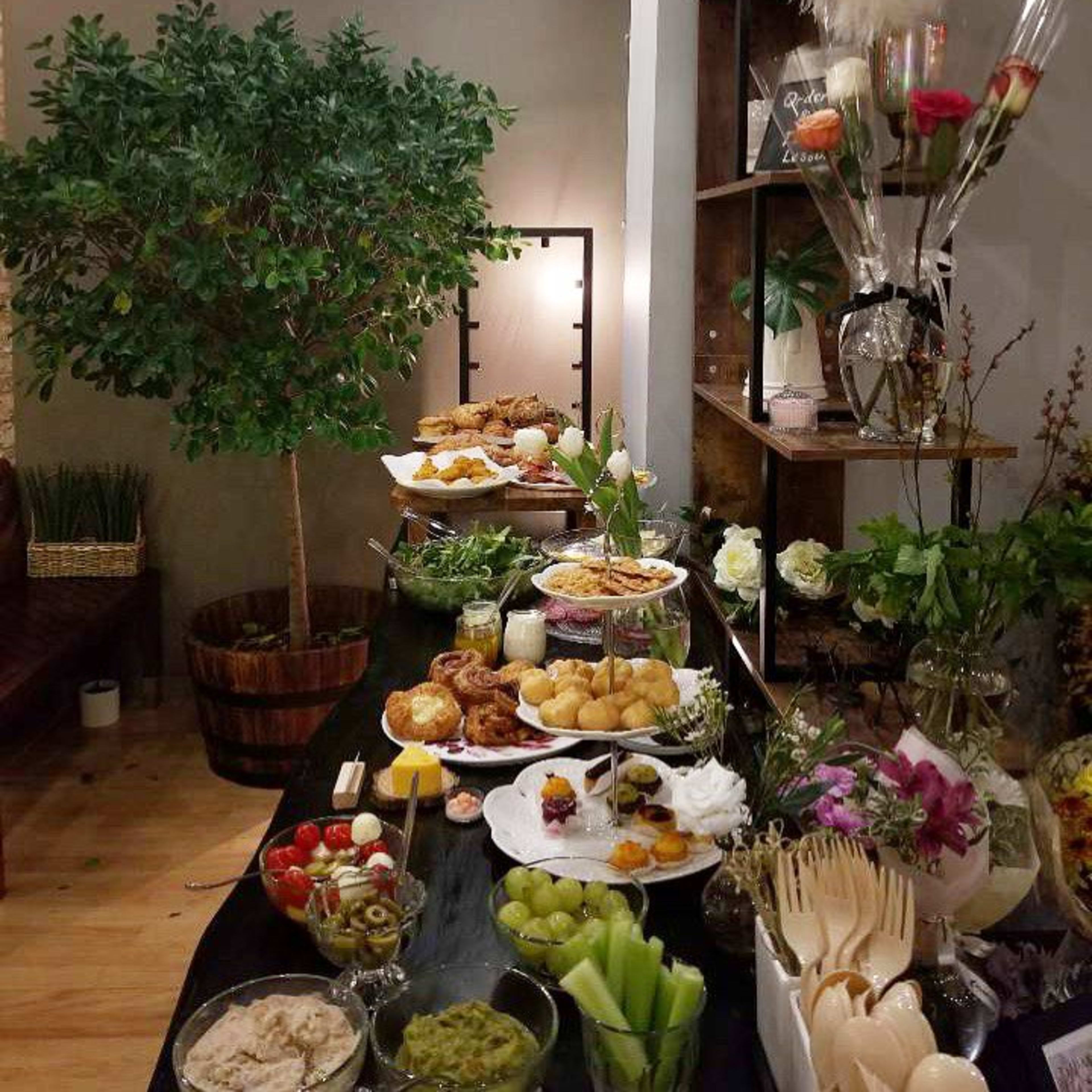 A table is set with various food platters, including fruits, pastries, and dips, alongside decorative plants and arrangements.