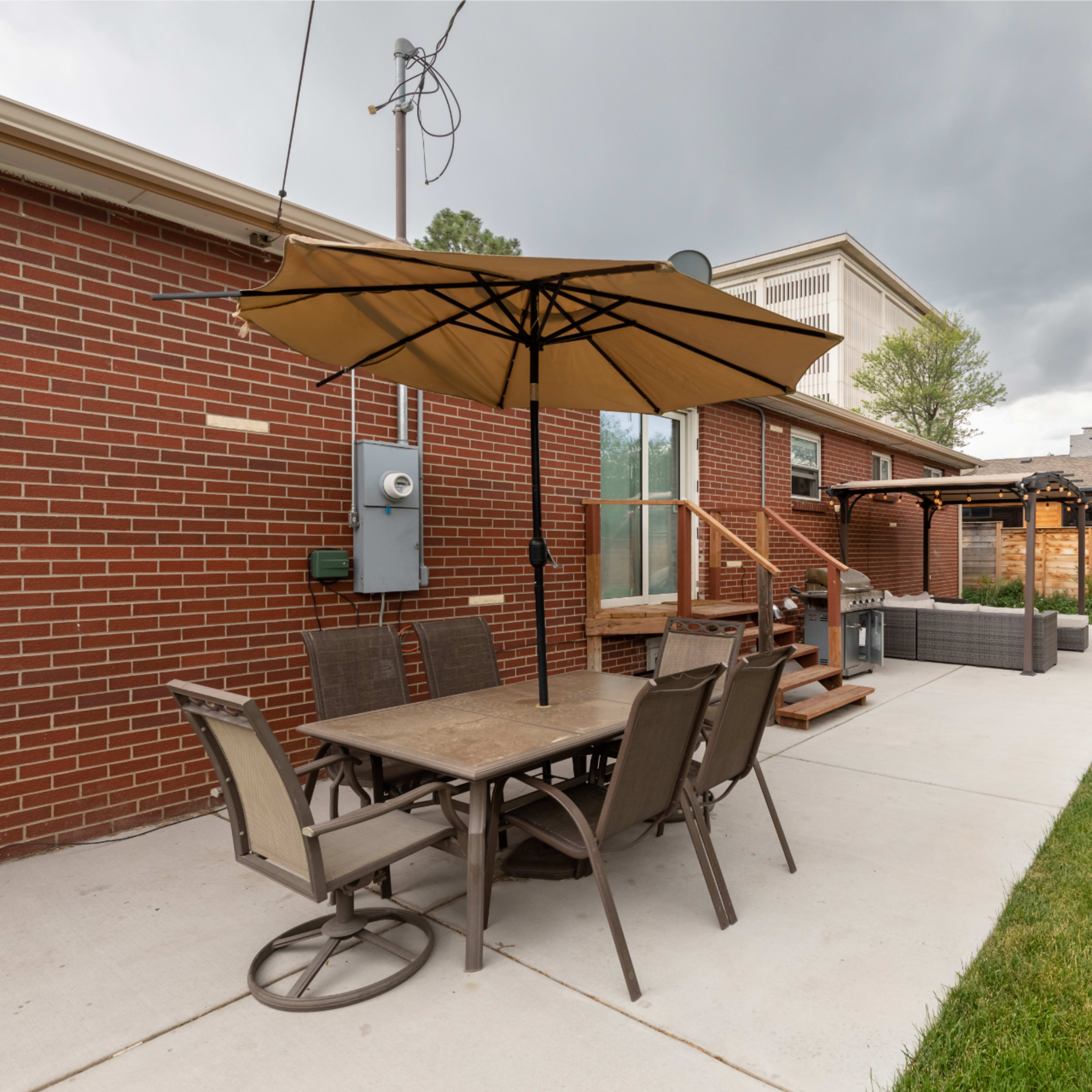 A patio area features a table with chairs and a large umbrella, adjacent to a brick house, with a grassy lawn and a wooden fence in the background.