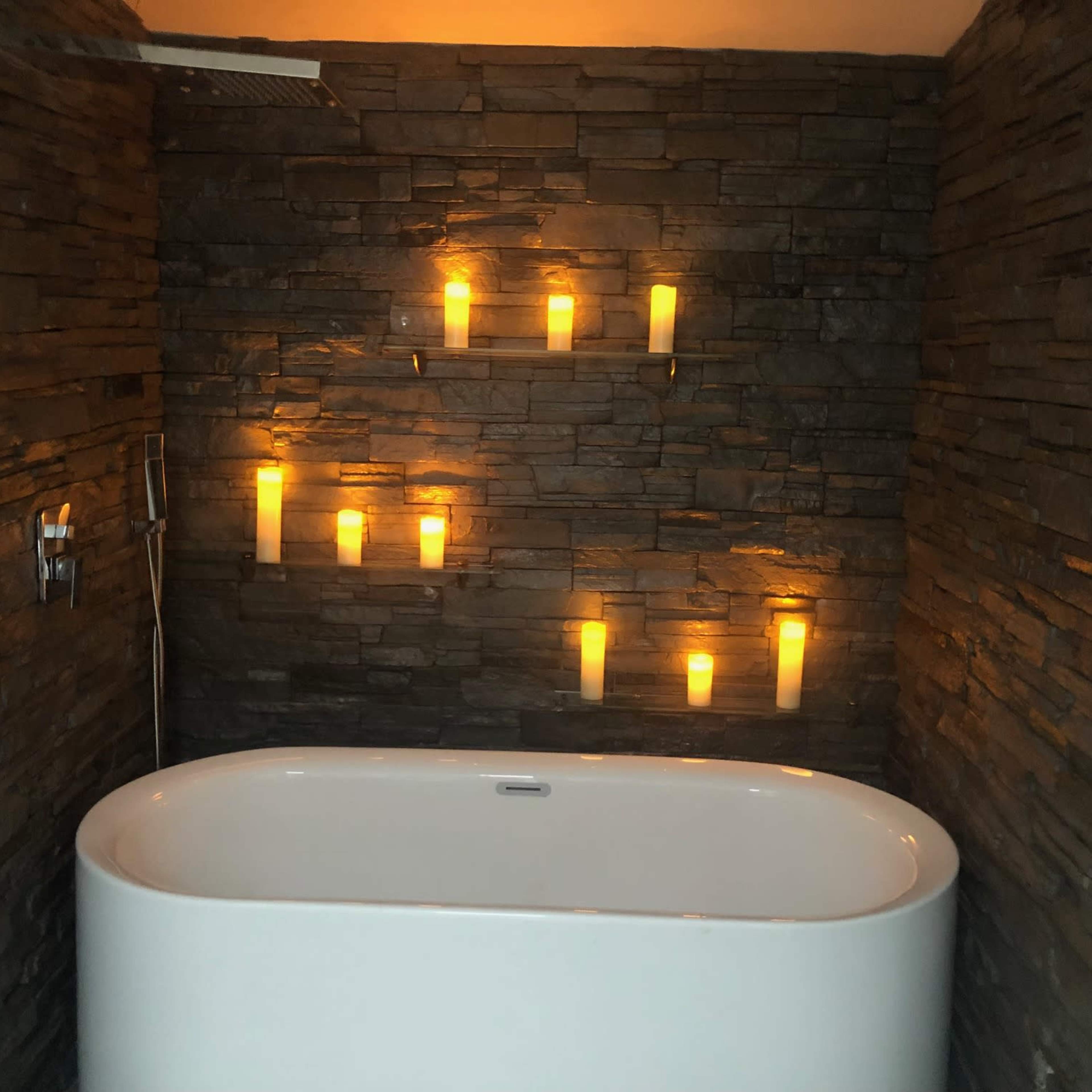 A modern bathtub is positioned against a stone wall, illuminated by several wall-mounted candles.