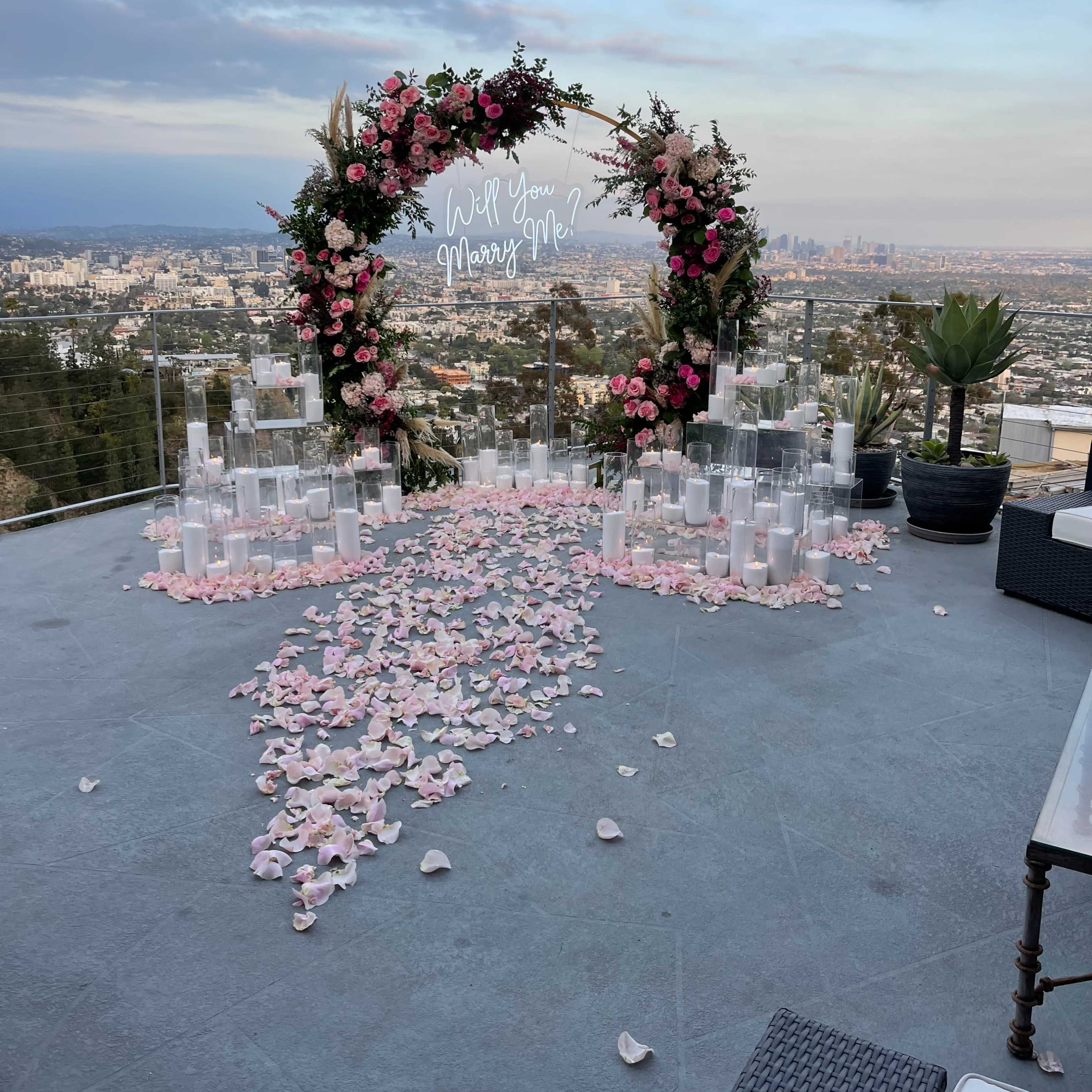 A decorative arch adorned with flowers and an illuminated sign stands on a terrace overlooking a city skyline, surrounded by a path made of rose petals and candles.