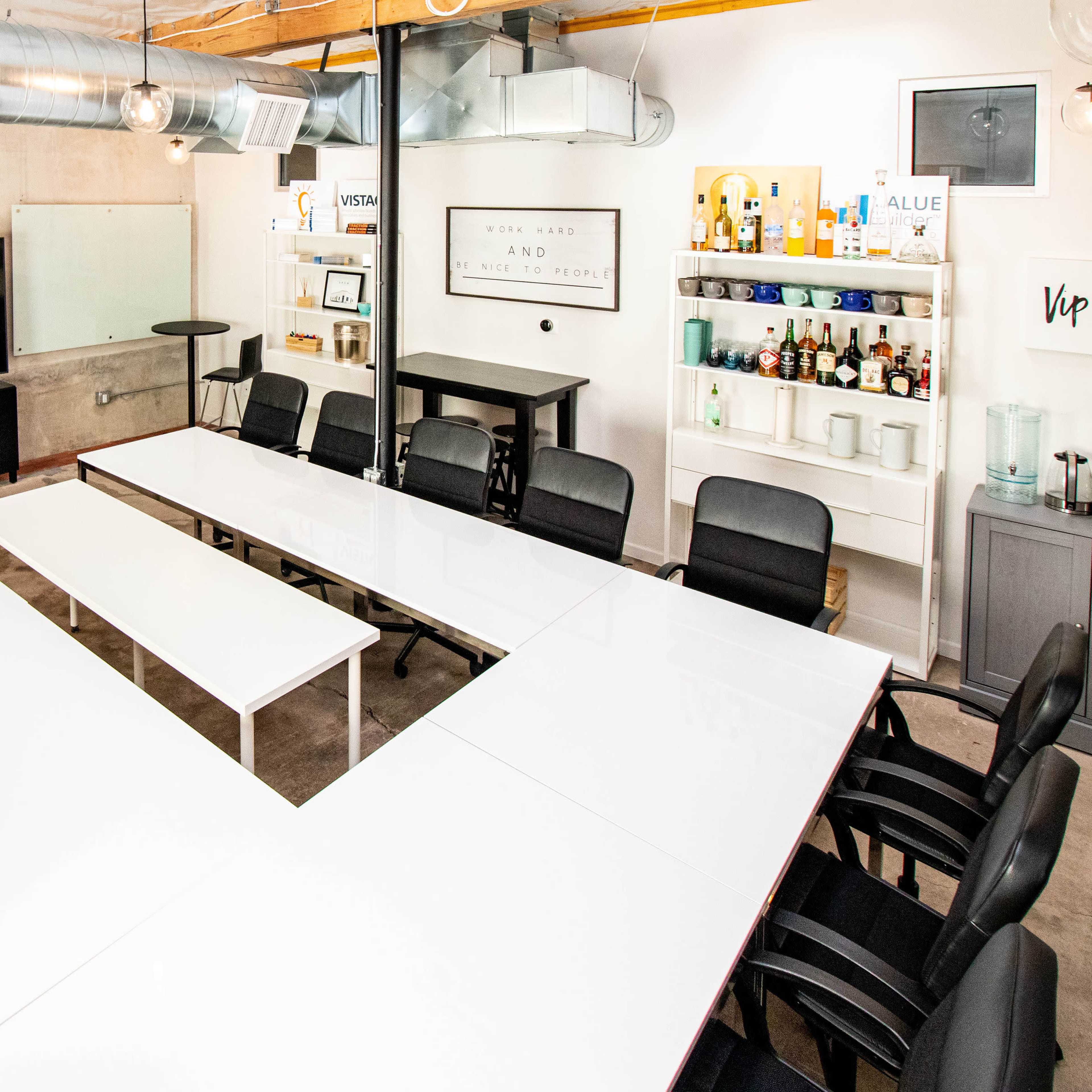 A modern conference room features a large white table arranged in a U-shape, surrounded by black chairs, with a TV display, whiteboards, and a bar cart in the background.