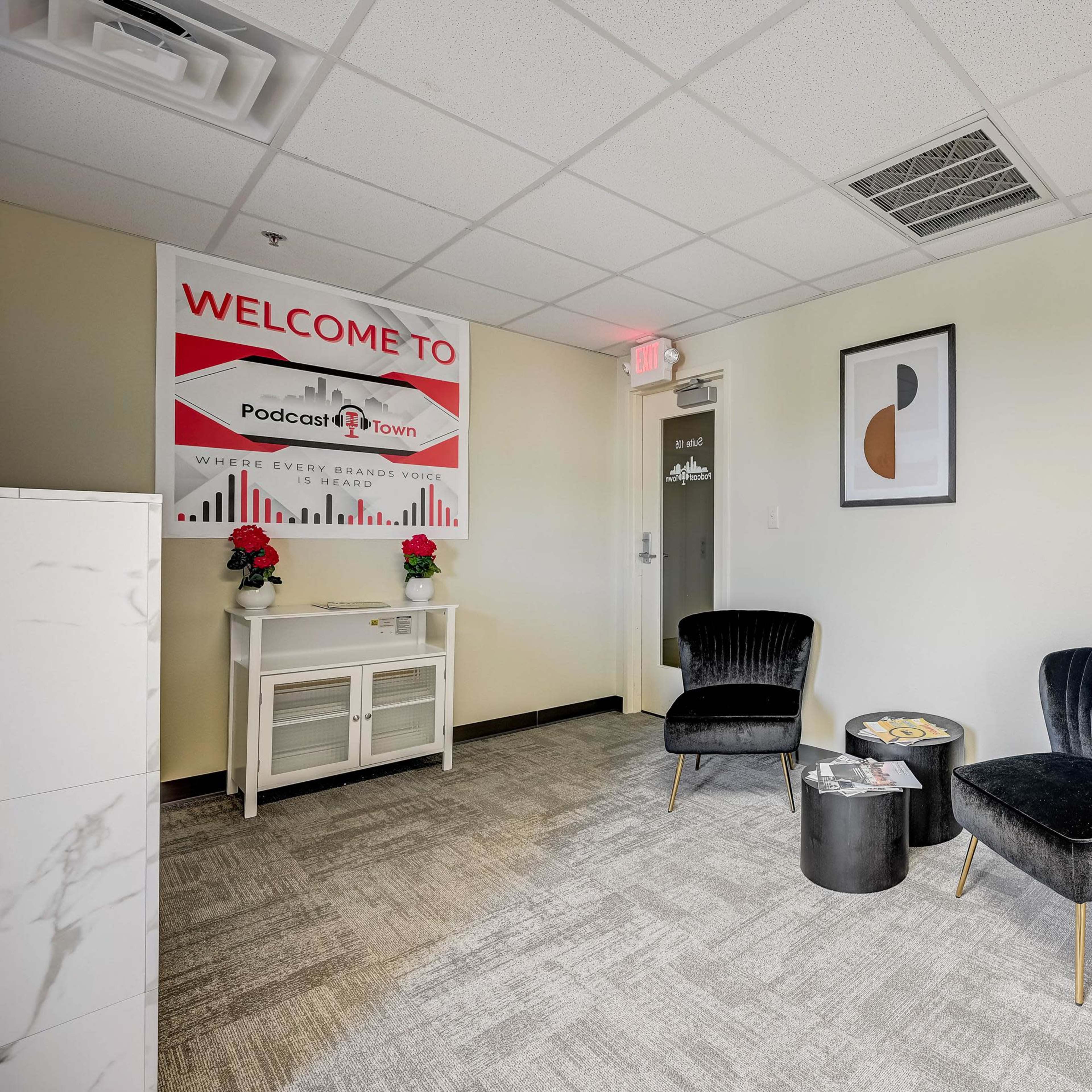 A modern office space featuring two black chairs, a small black table, a red fridge, and a sign that says "Welcome to PodcastTown."