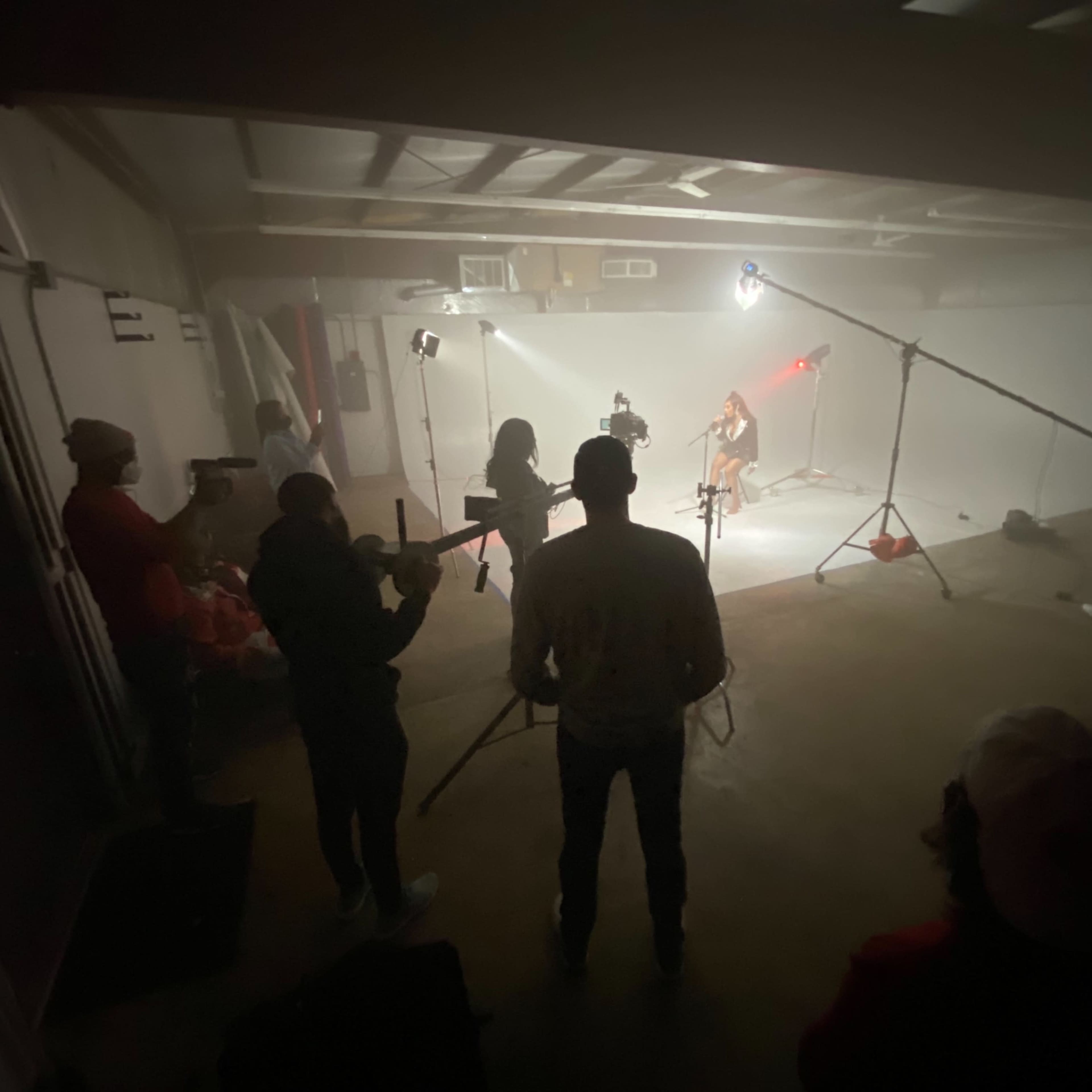 A group of individuals stands in a dimly lit room, observing a subject illuminated by lights on a set.