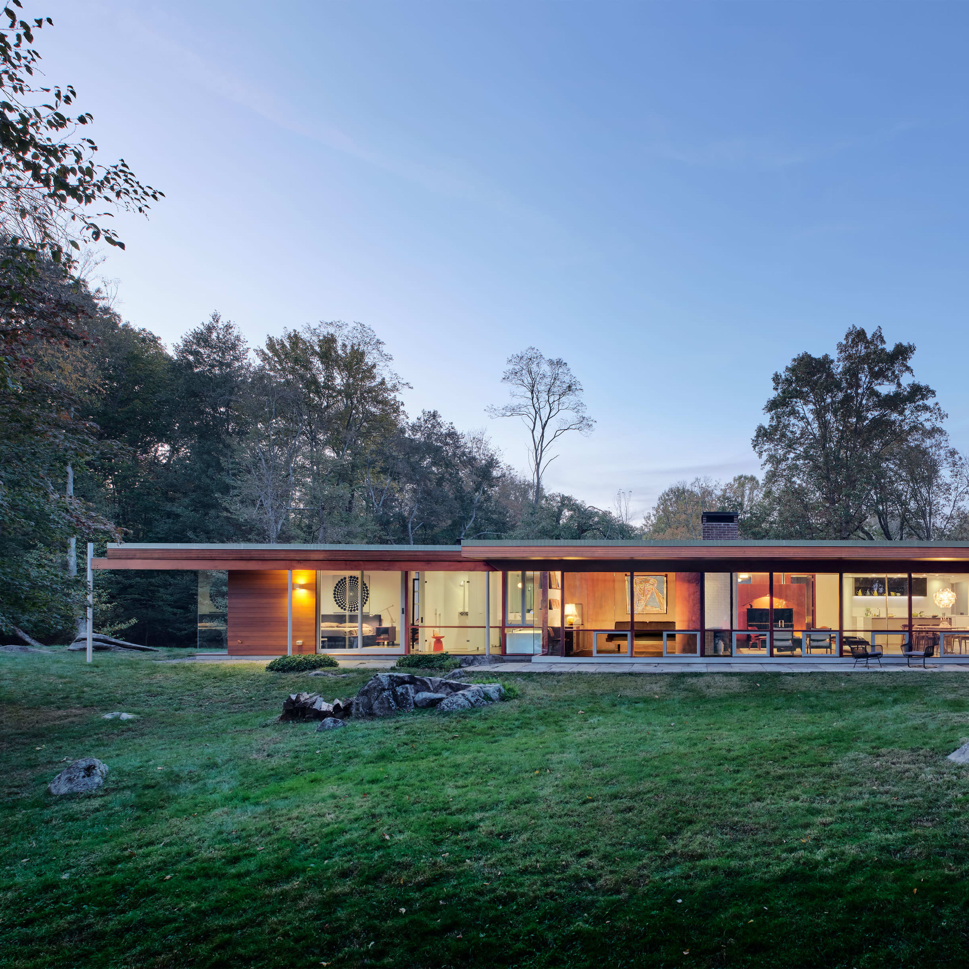 A single-story modern house with large glass windows is surrounded by trees and a grassy lawn.