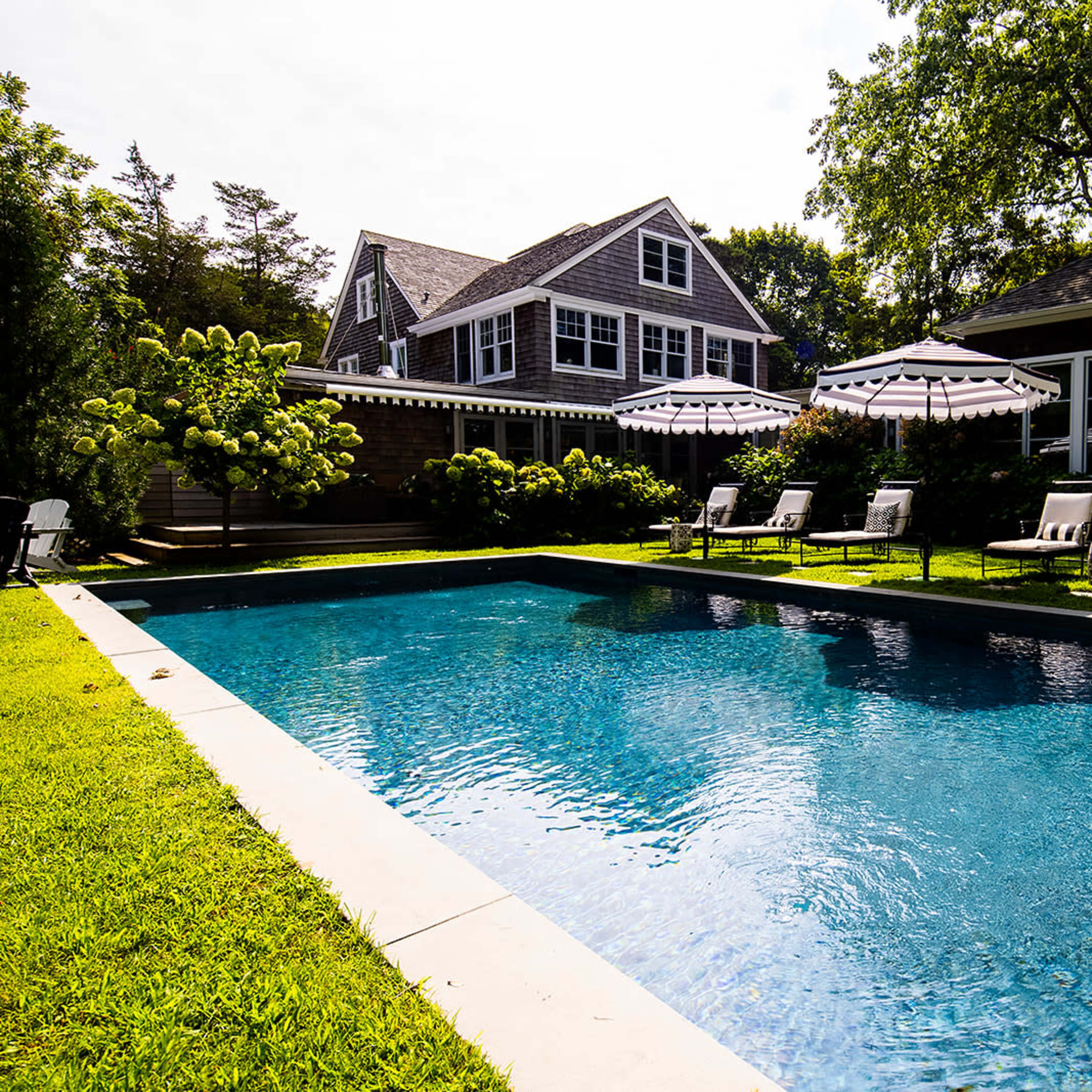 A well-maintained swimming pool surrounded by lounge chairs and a large house in the background.