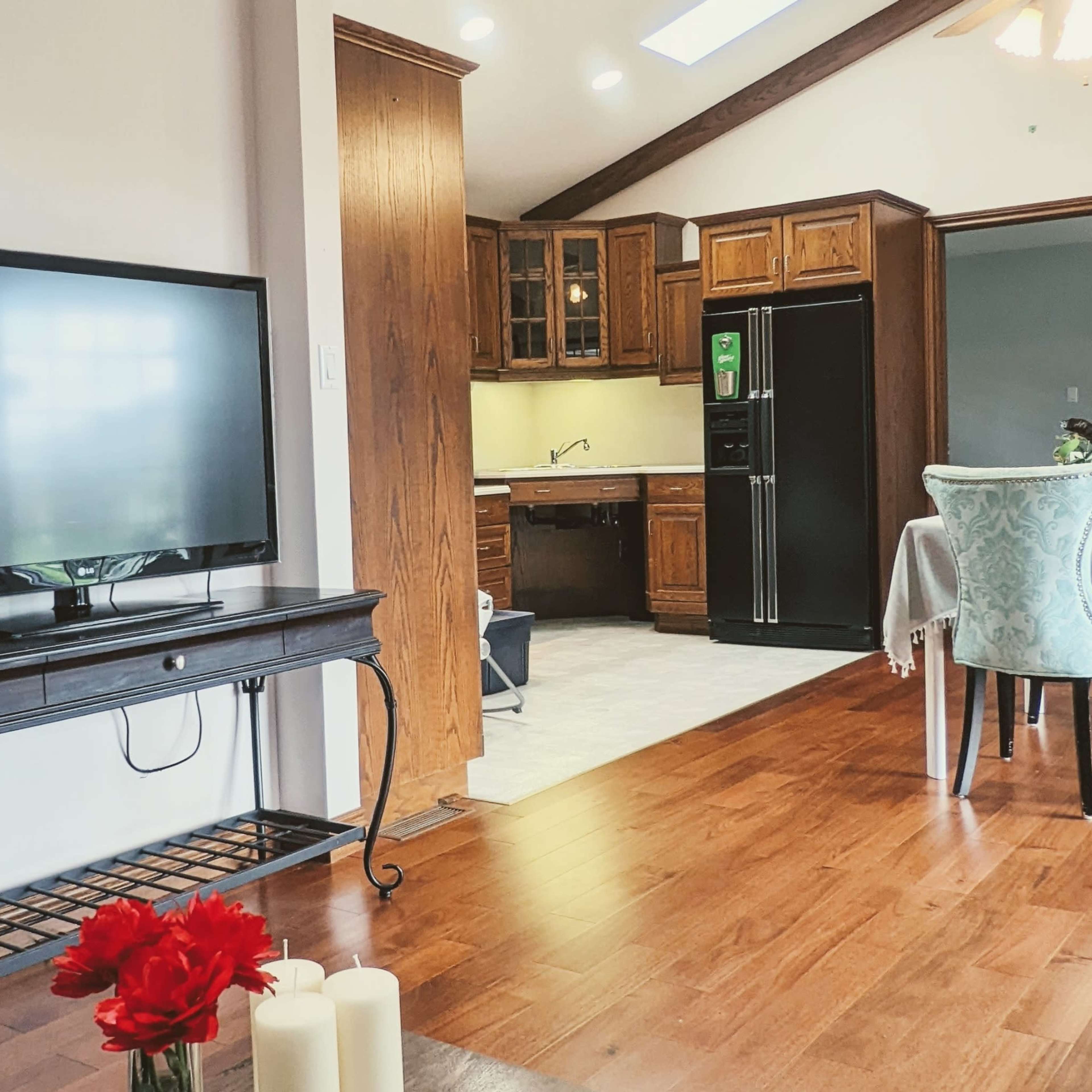 A living area with a television on a stand, looking into a kitchen with wooden cabinetry and a dining table set nearby.