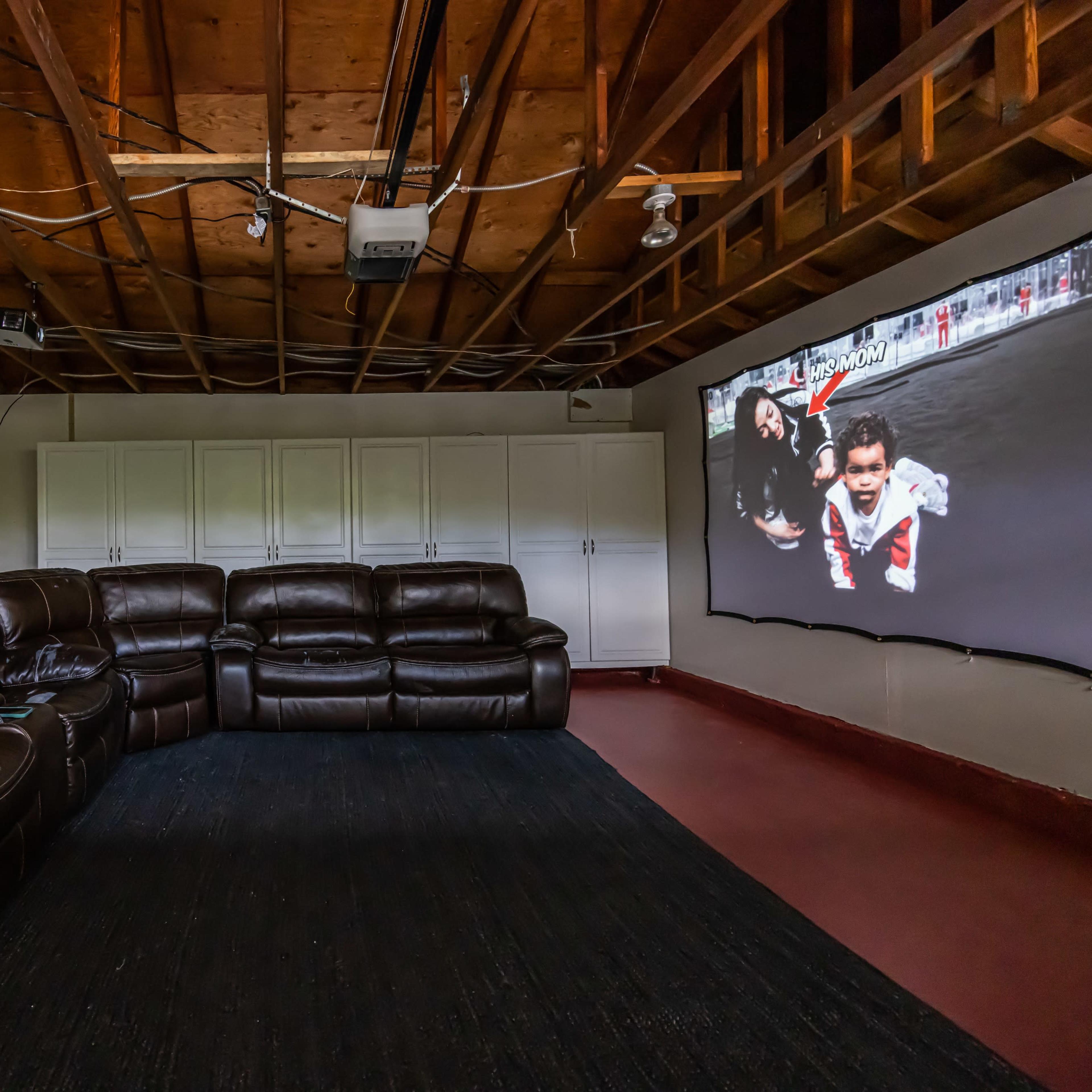 A home theater setup with a large screen displaying a sports scene, surrounded by a dark sectional sofa and white storage cabinets.