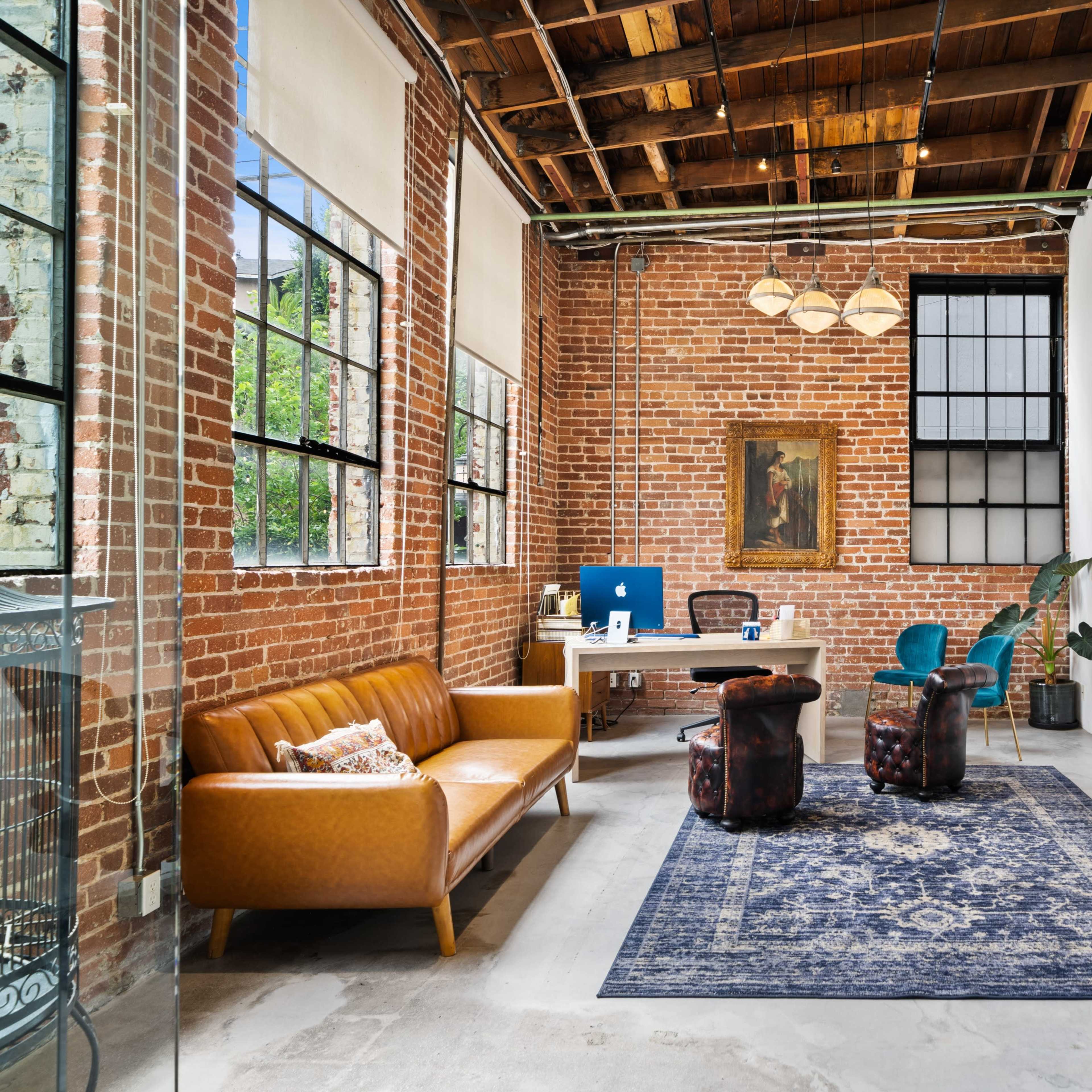 A modern office space featuring exposed brick walls, wooden beams, a large window, and various furnishings including a brown leather sofa and blue chairs.
