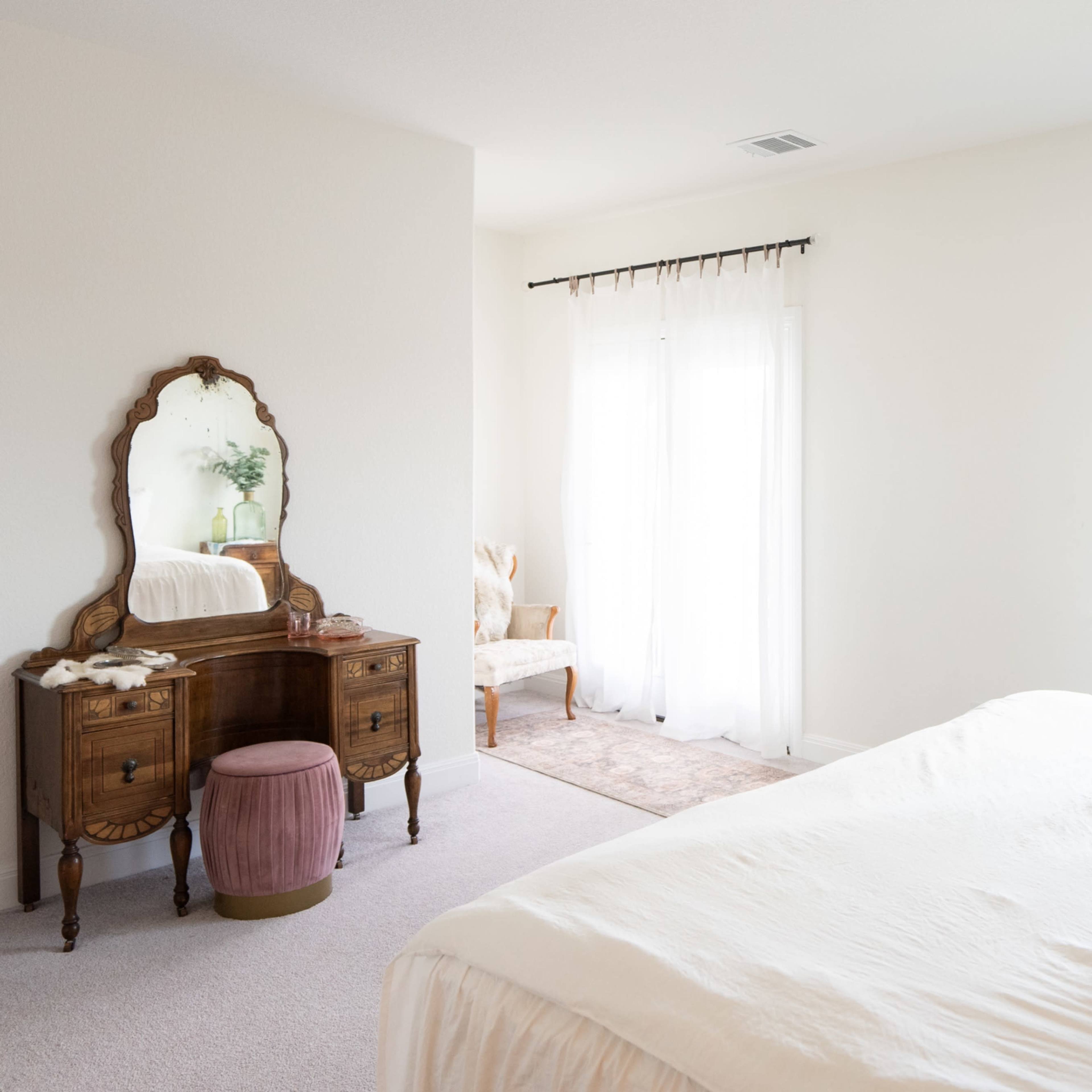 A bright bedroom features a bed with white linens, a wooden vanity with a mirror, and an arched window letting in natural light.