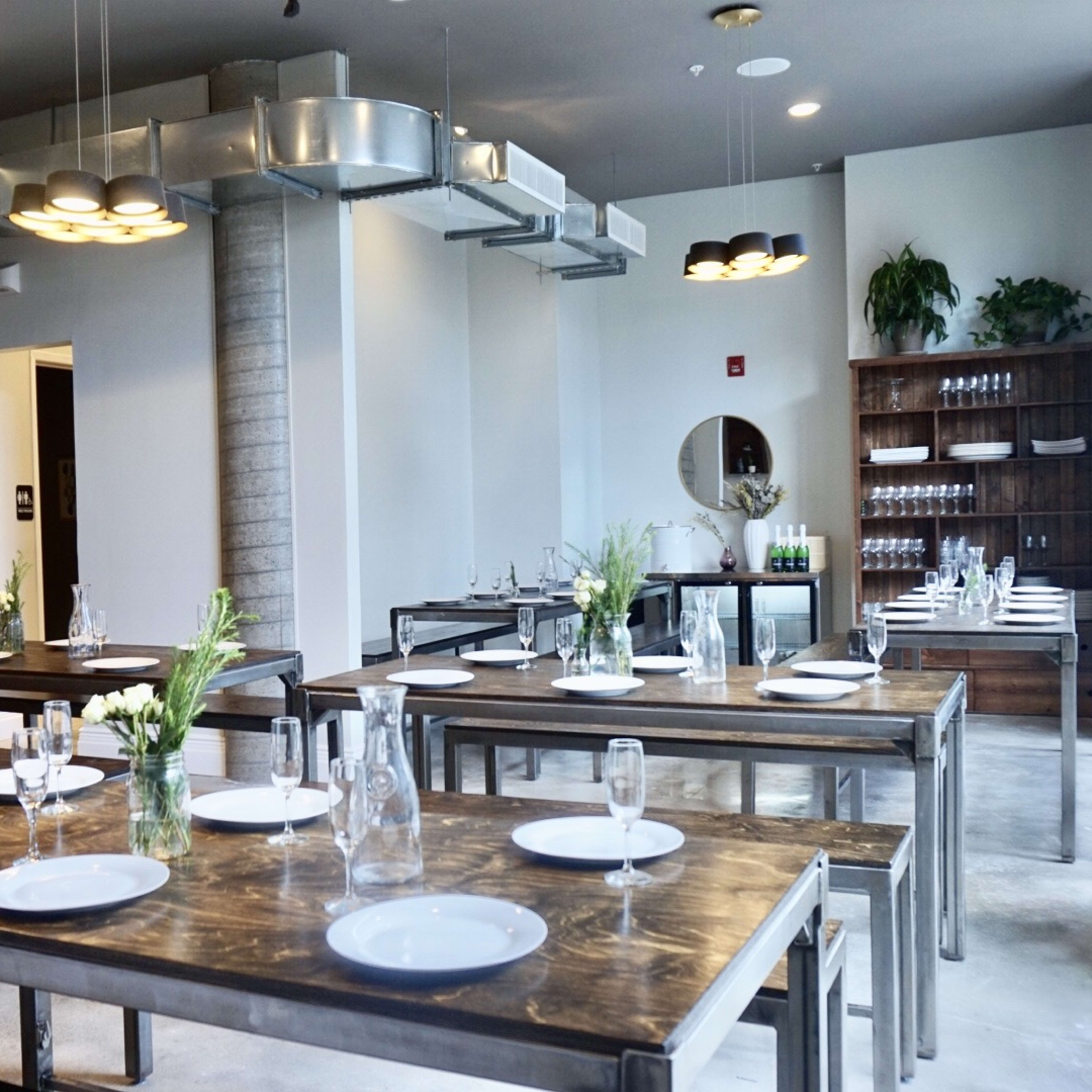 A modern dining area with several long wooden tables, each set with white plates and vases of flowers, and a wall lined with shelves holding glassware.