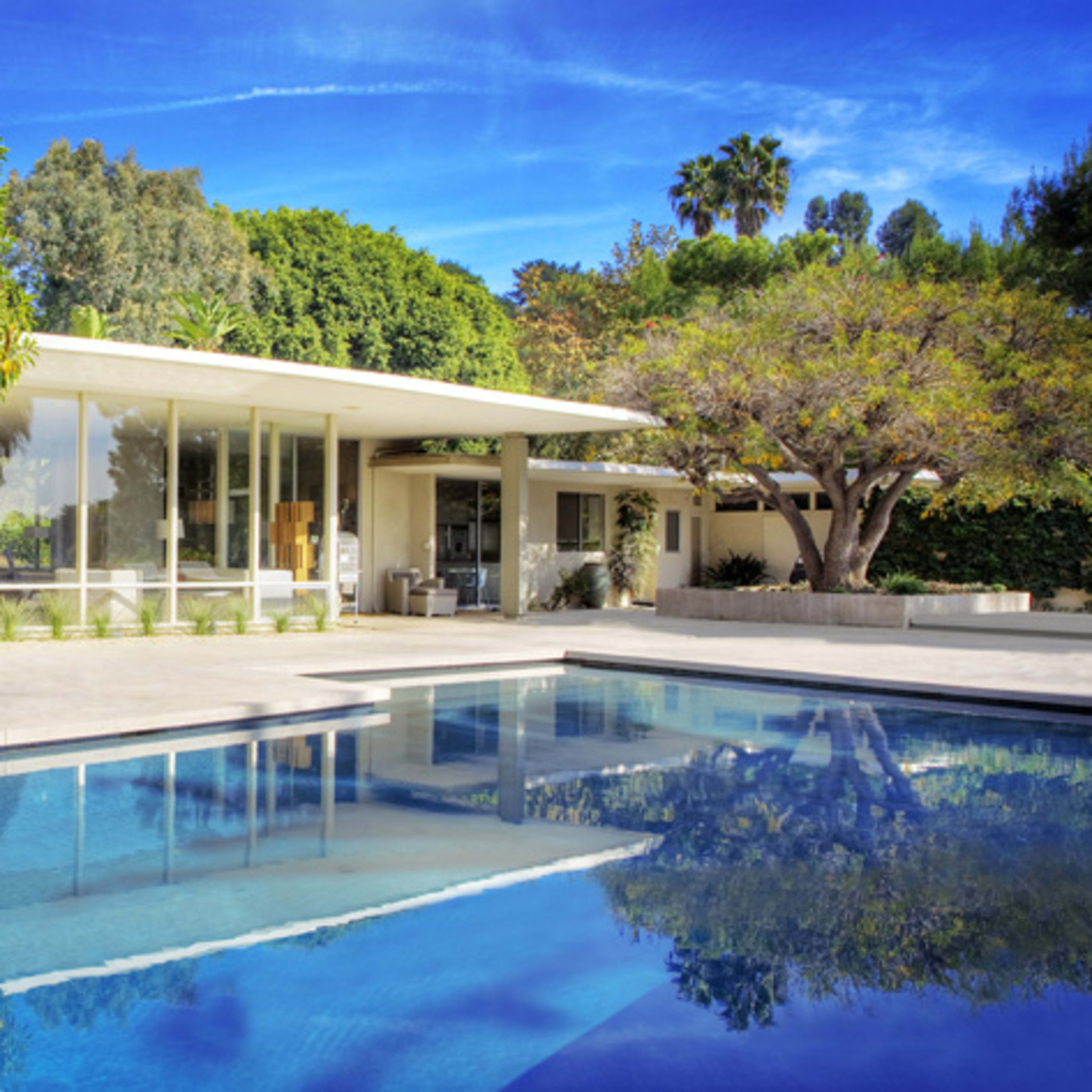 A modern house with large glass windows surrounded by trees and a reflective swimming pool in the foreground.