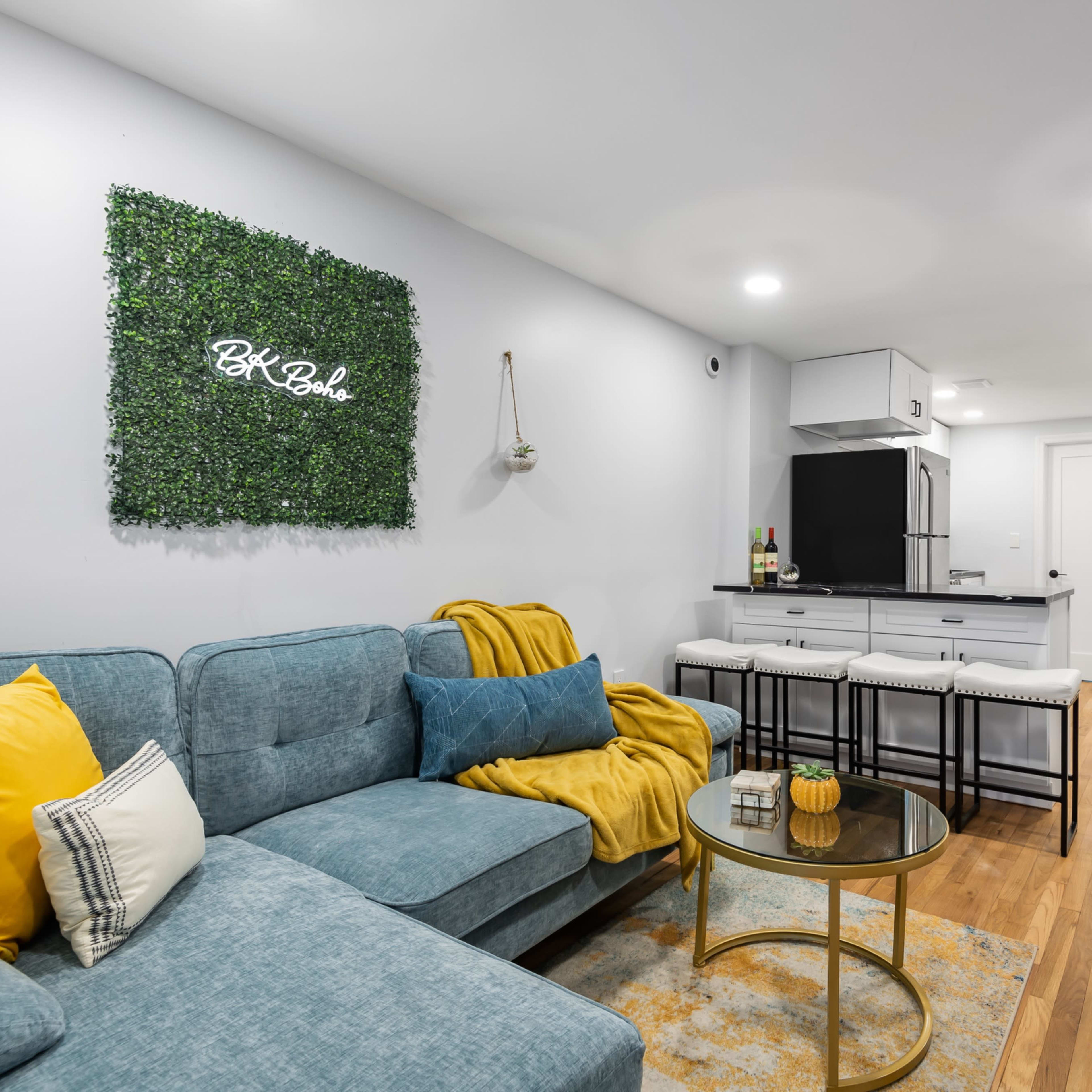 A modern living area with a light gray wall, a teal sectional sofa, a round glass coffee table, and a kitchenette in the background, featuring a bar area with stools.
