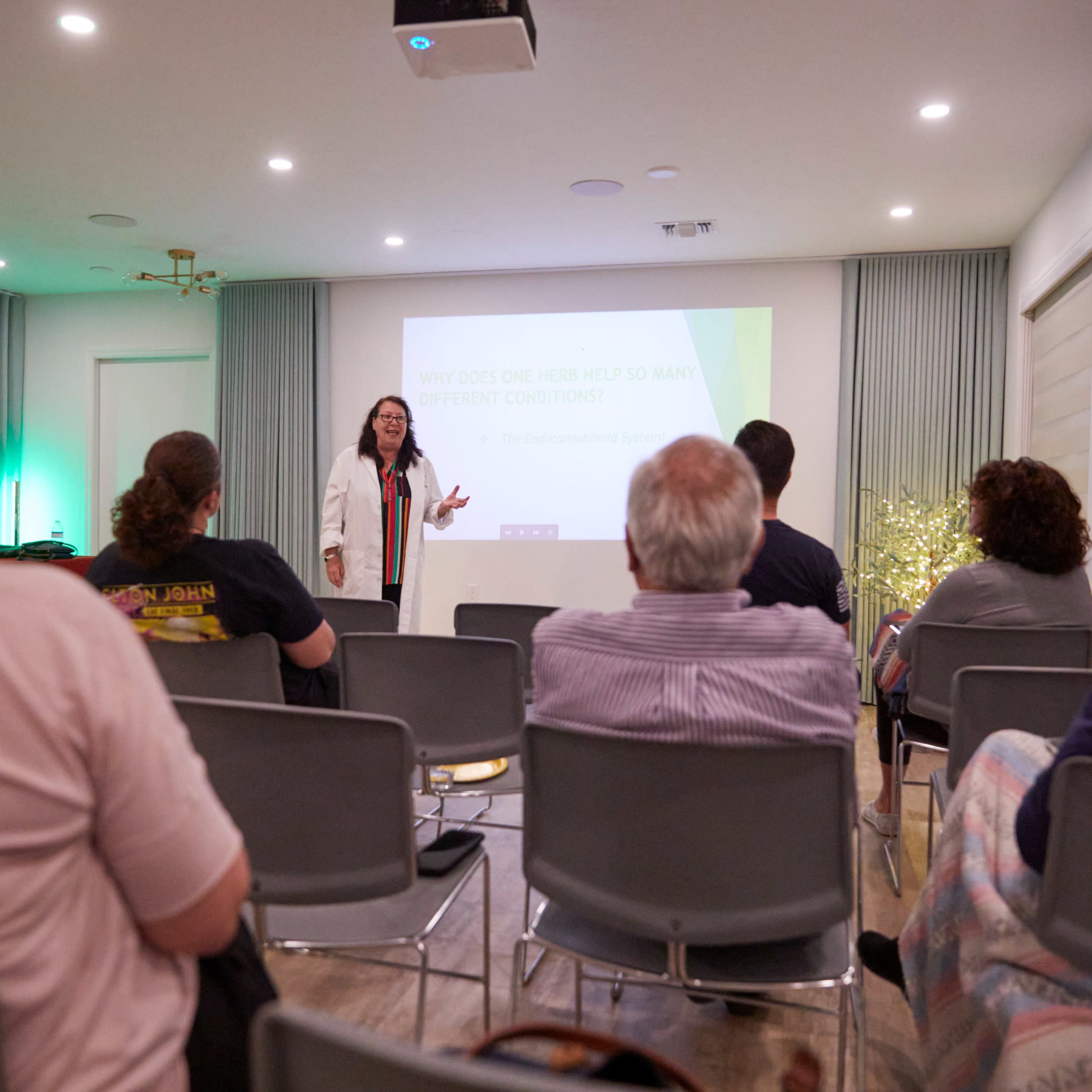 A presenter stands in front of an audience seated in a room with a projection screen, delivering a lecture on the benefits of a certain treatment for various conditions.