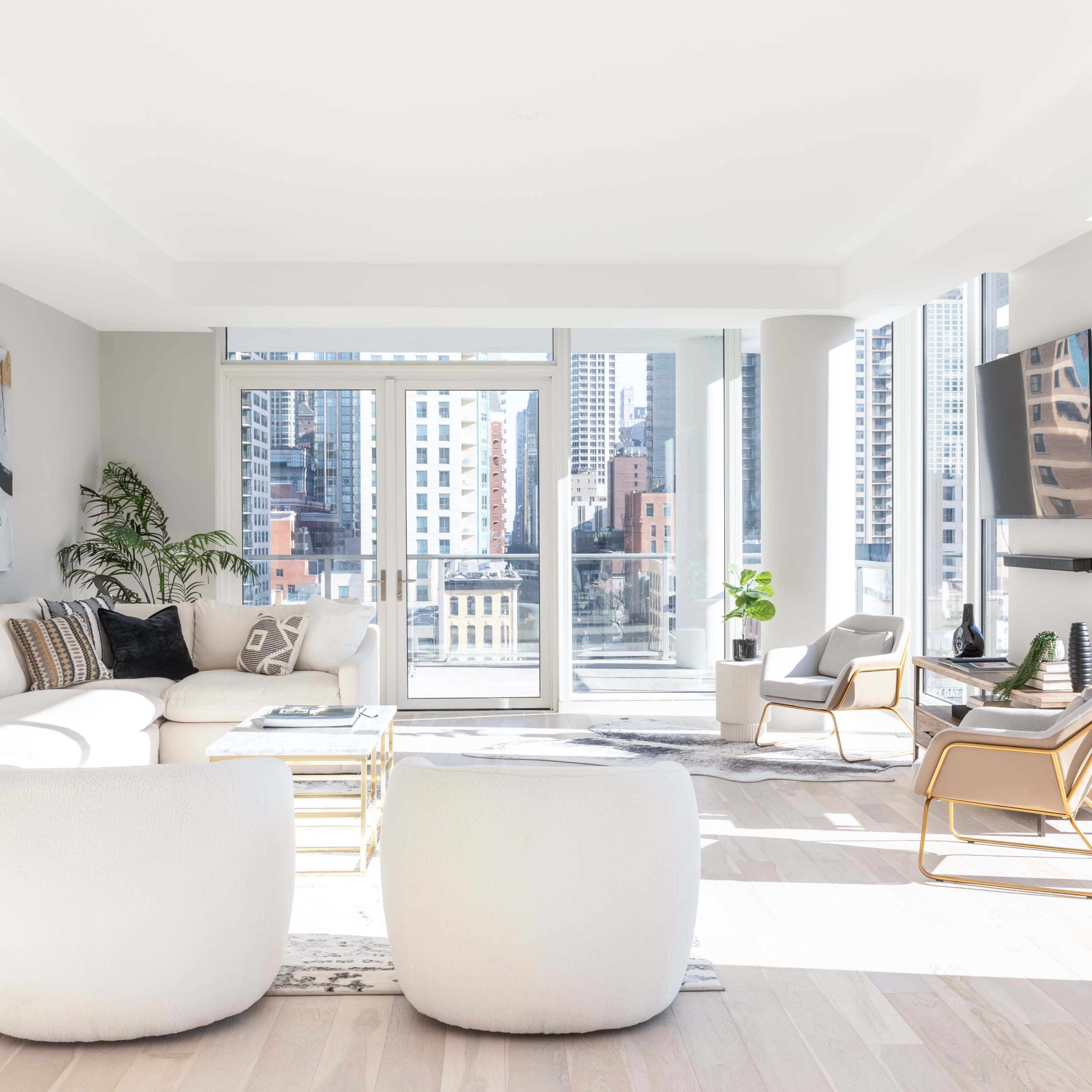 A modern, bright living room with large windows offering a view of a city skyline, featuring a light-colored sofa, two round stools, a coffee table, and a television mounted on the wall.
