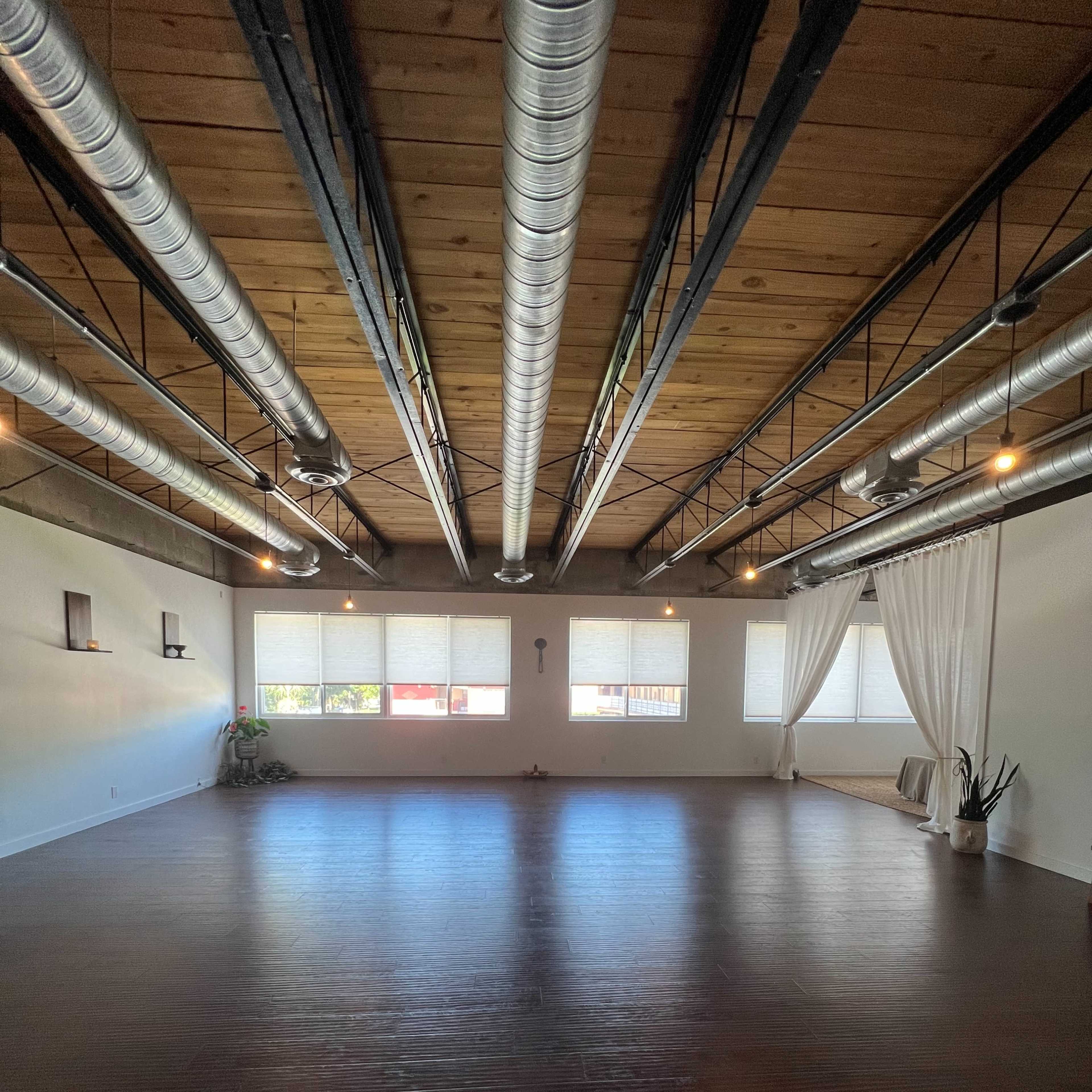 An empty indoor space with a wooden floor, large windows, and exposed metal ductwork on the ceiling.