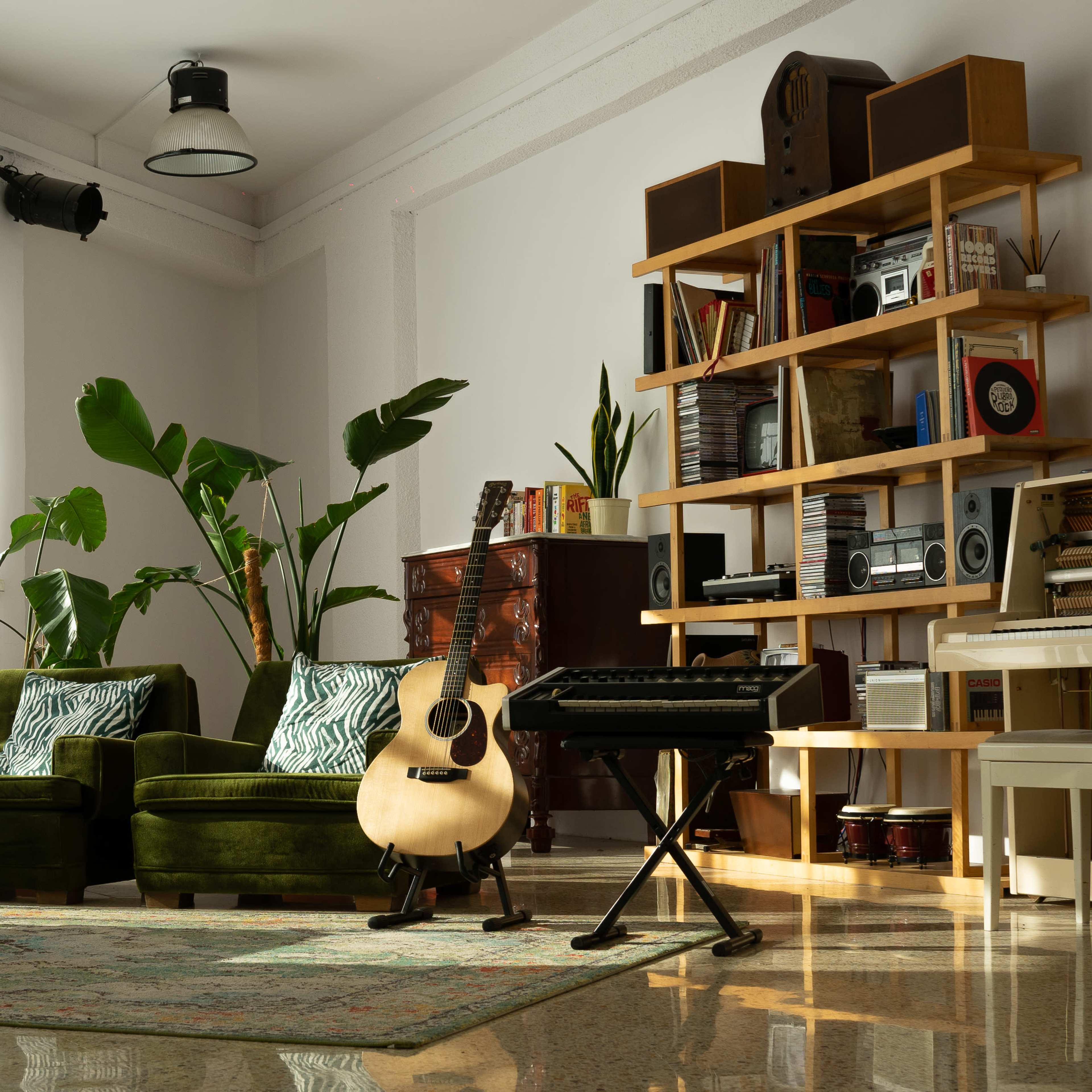 A cozy living room featuring green upholstered chairs, a wooden bookshelf filled with books and electronics, a guitar on a stand, and several indoor plants.