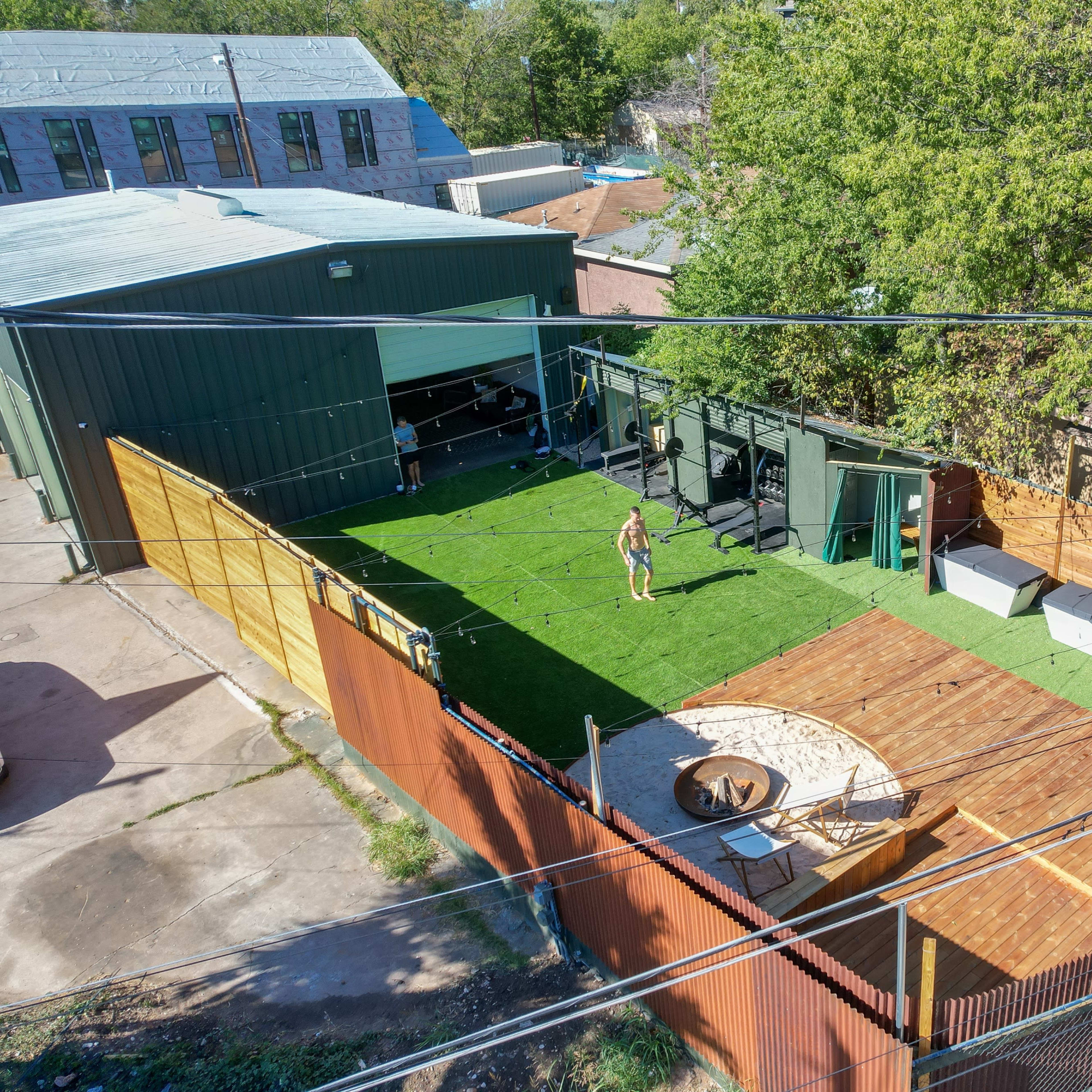 An outdoor space featuring green grass, wooden decks, seating areas, and a circular fire pit, surrounded by fences and buildings.