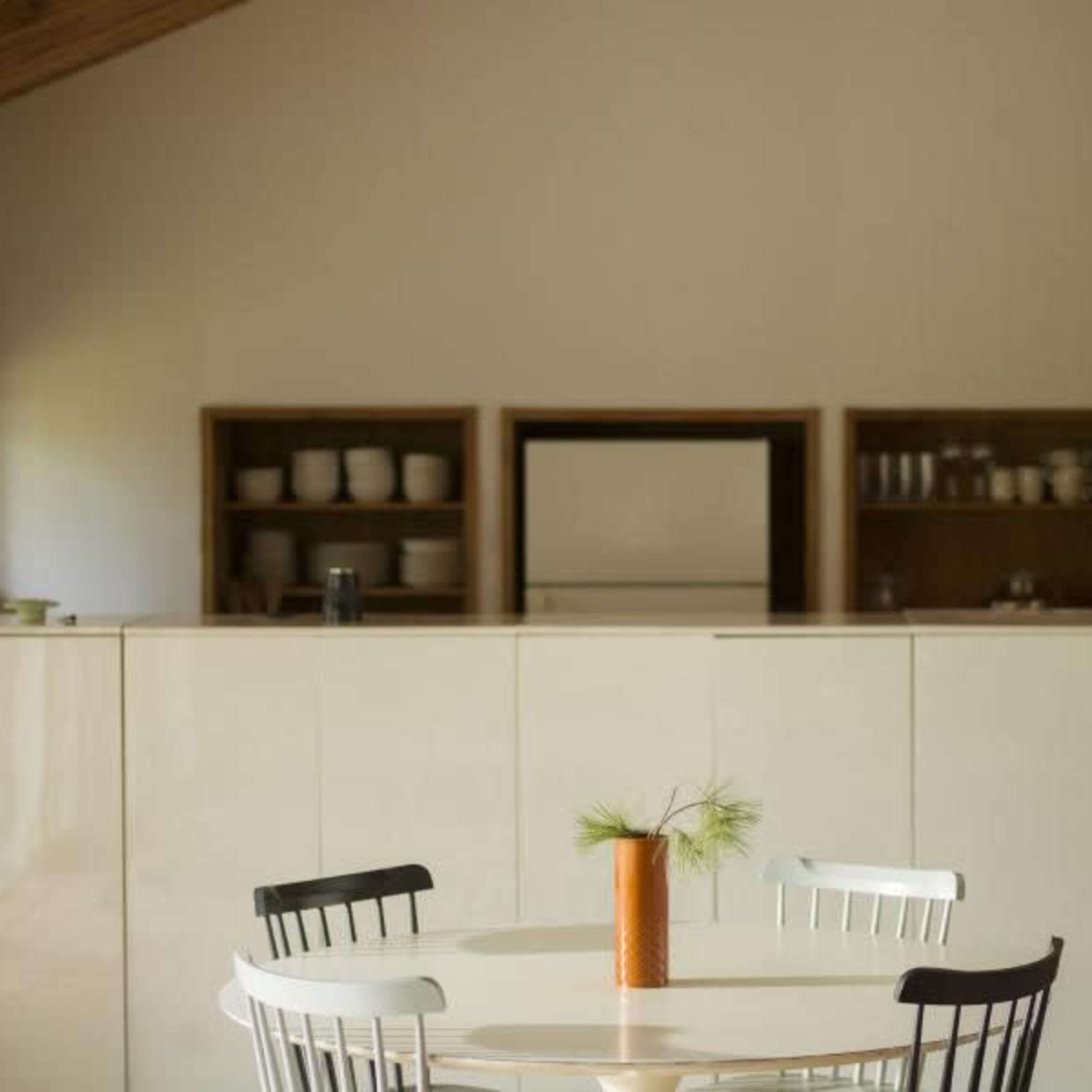 A round table with four chairs and a vase sits in the foreground of a minimalist kitchen featuring cabinetry and shelving in the background.