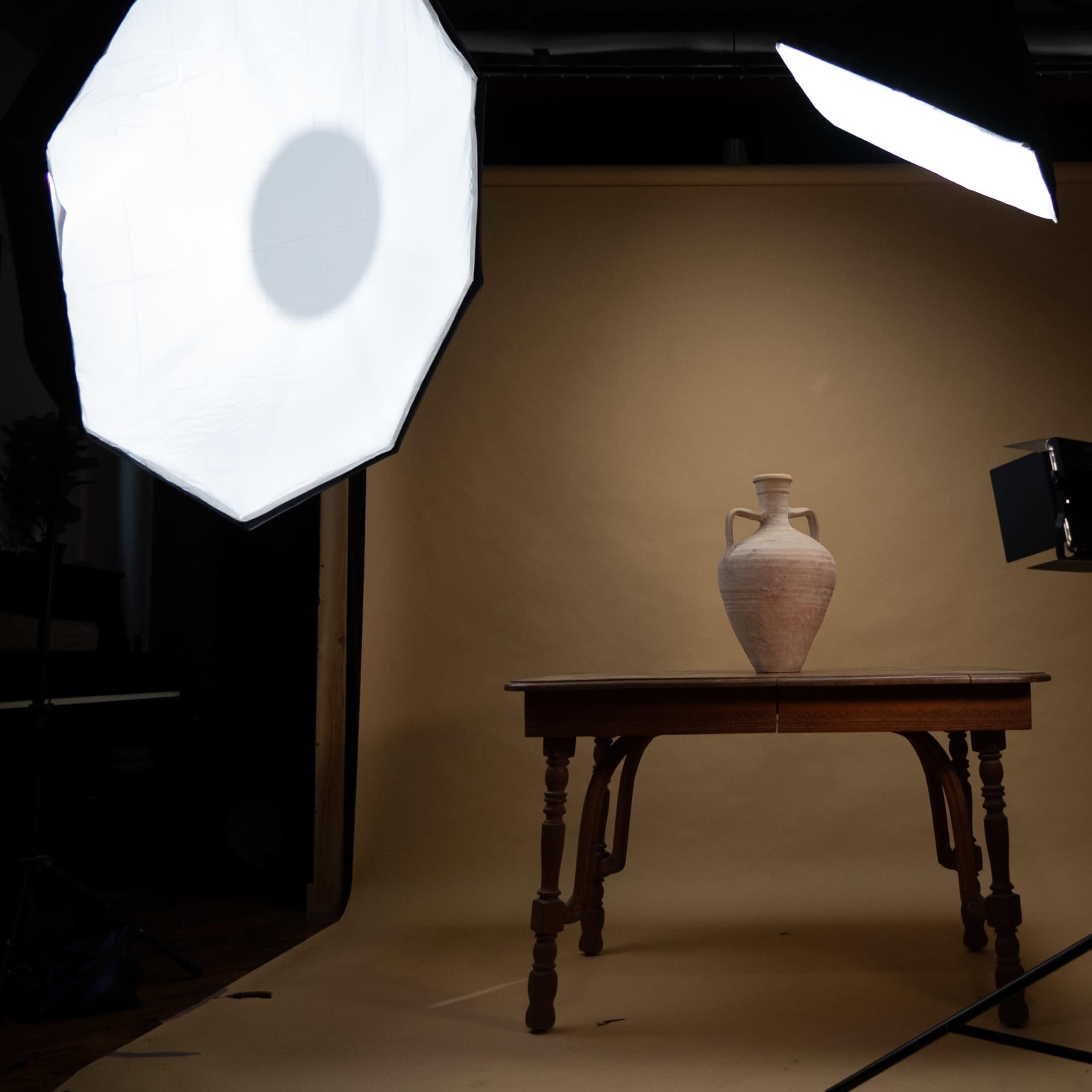 A pottery vase sits on a wooden table in a photography studio, illuminated by two soft light sources.