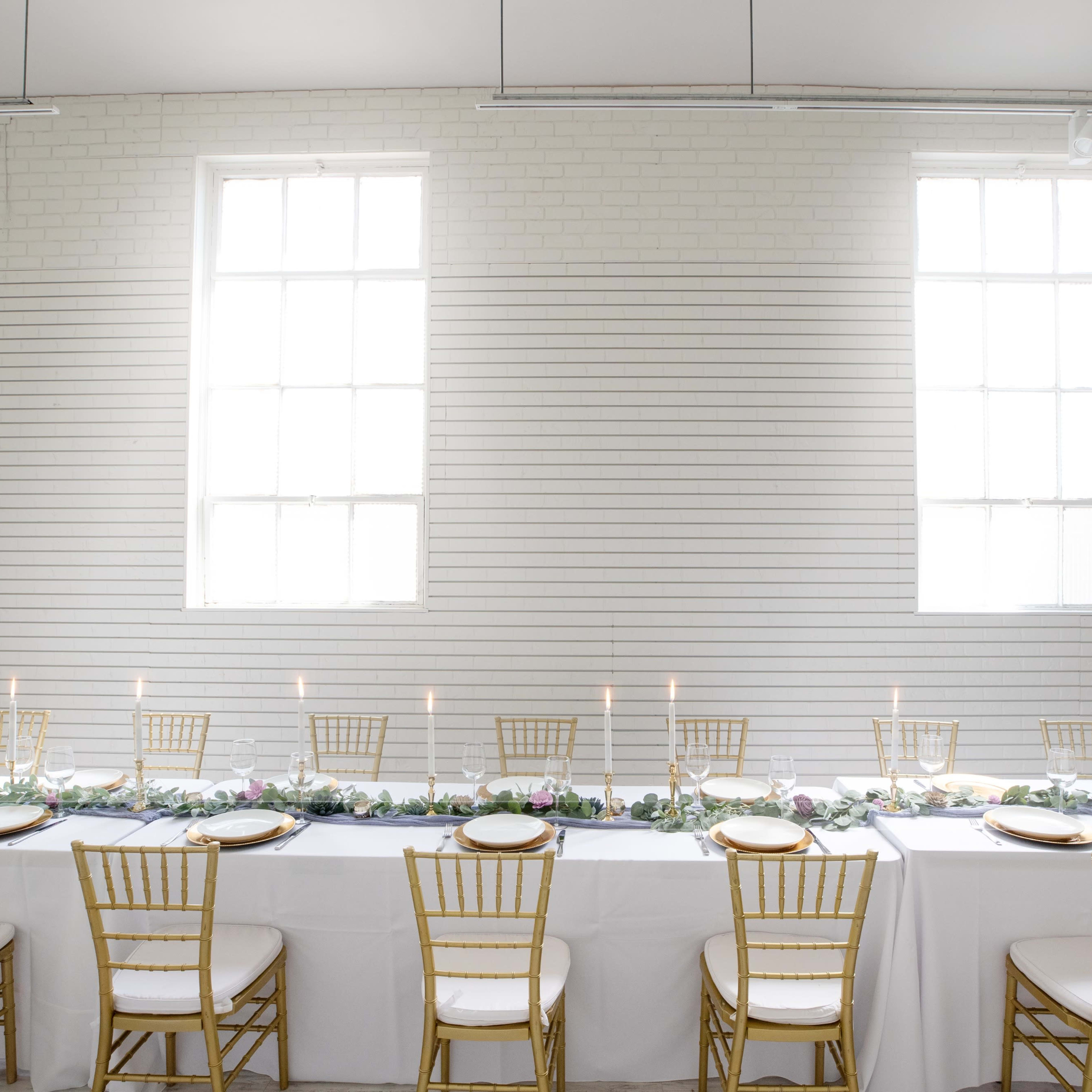 A long banquet table with white linens and gold chairs is set up against a wall with large windows, featuring a centerpiece of greenery and candles.
