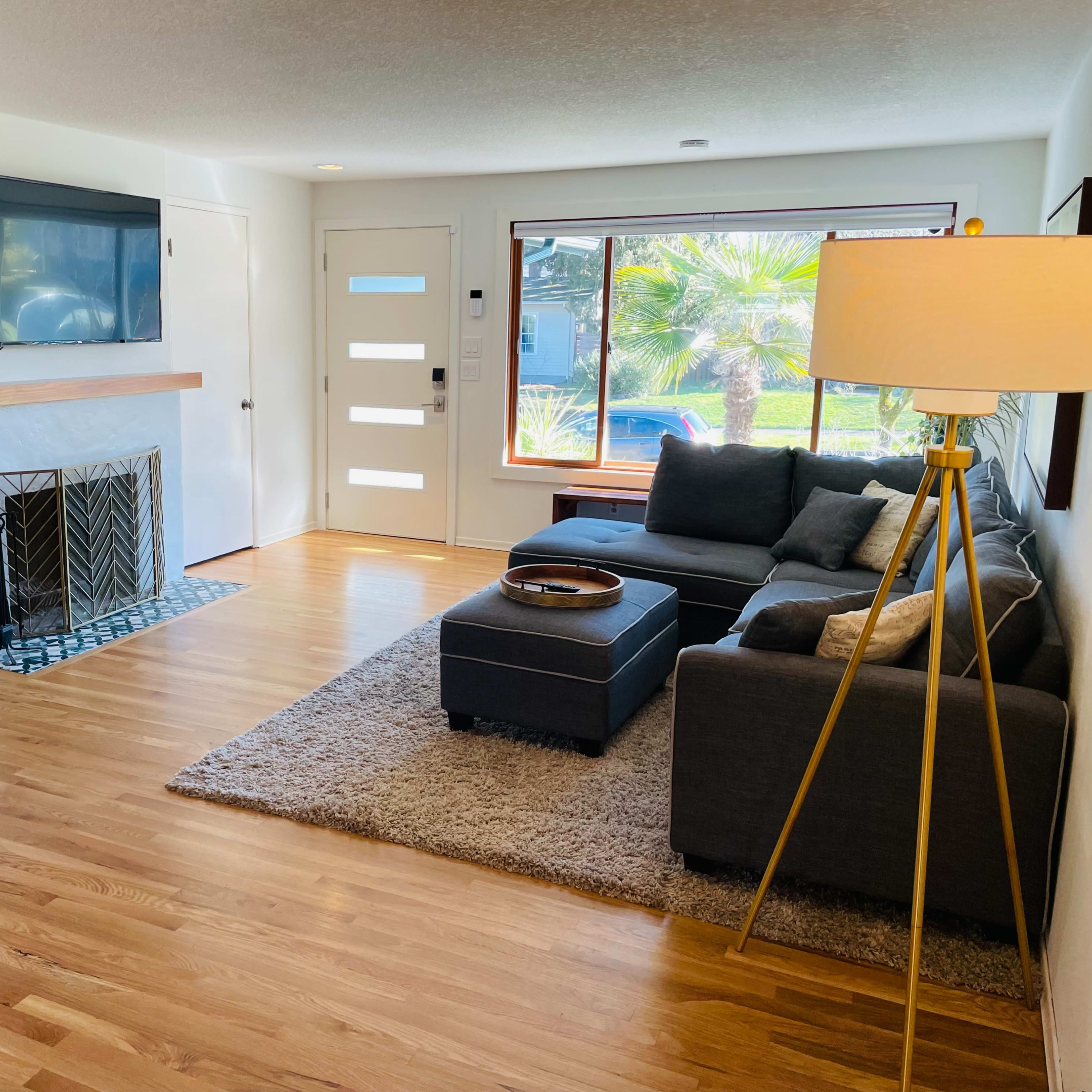 The living room features a gray sectional sofa, a coffee table, a flat-screen TV mounted on the wall, and a fireplace, with large windows overlooking a garden.