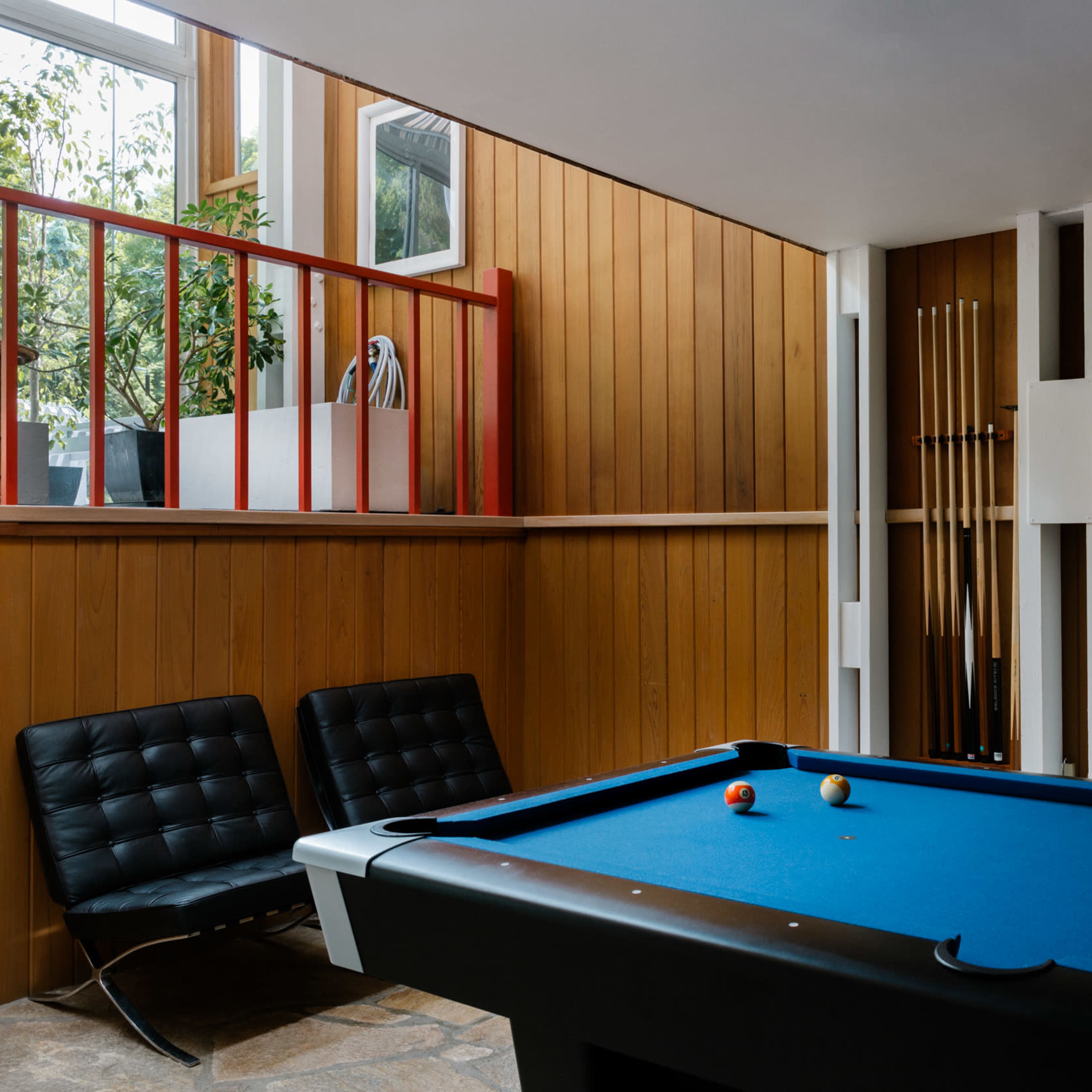 A room featuring a billiards table, two black lounge chairs, wooden paneling, and a staircase with potted plants in the background.
