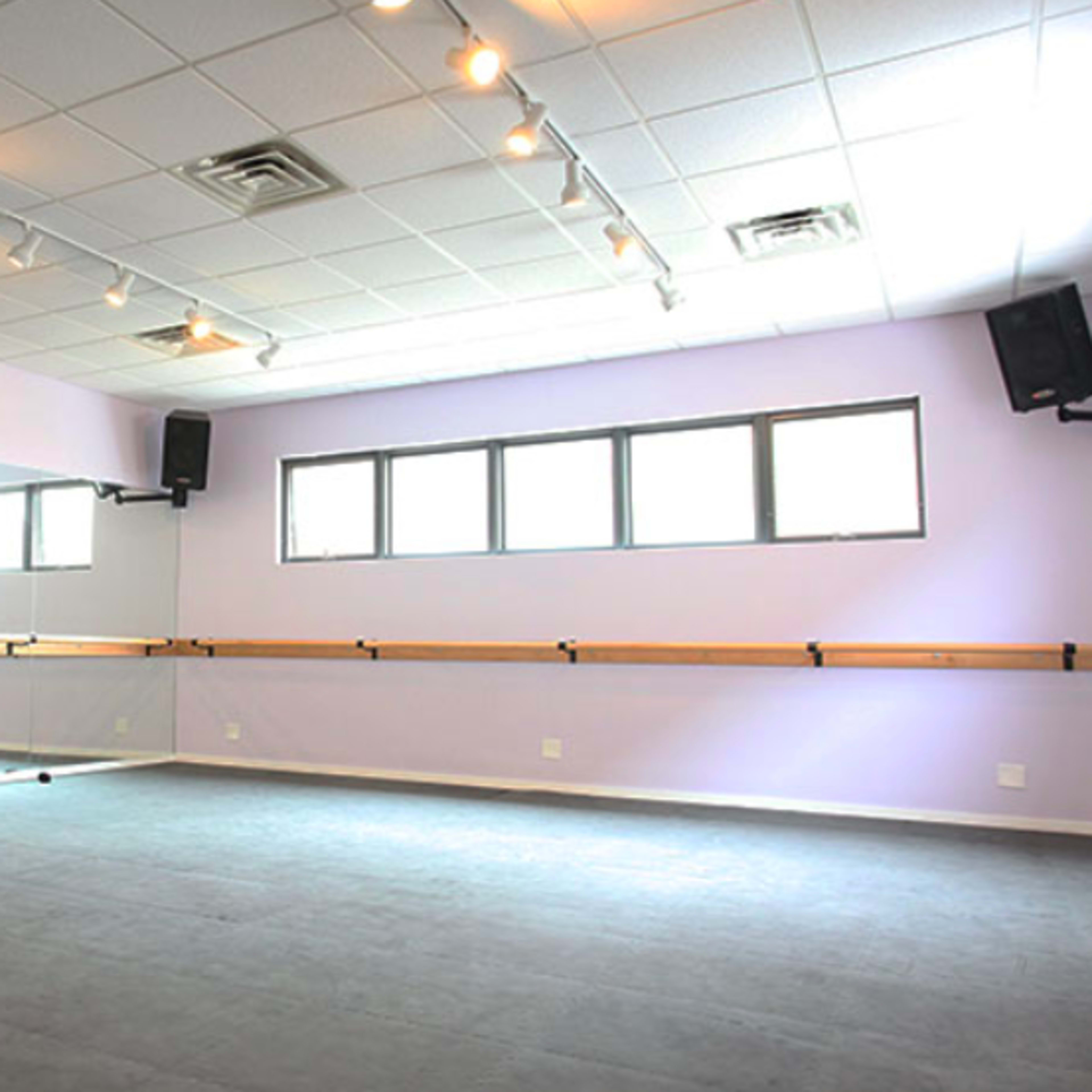 A dance studio with a mirrored wall, barre, and soft gray flooring under bright overhead lights.