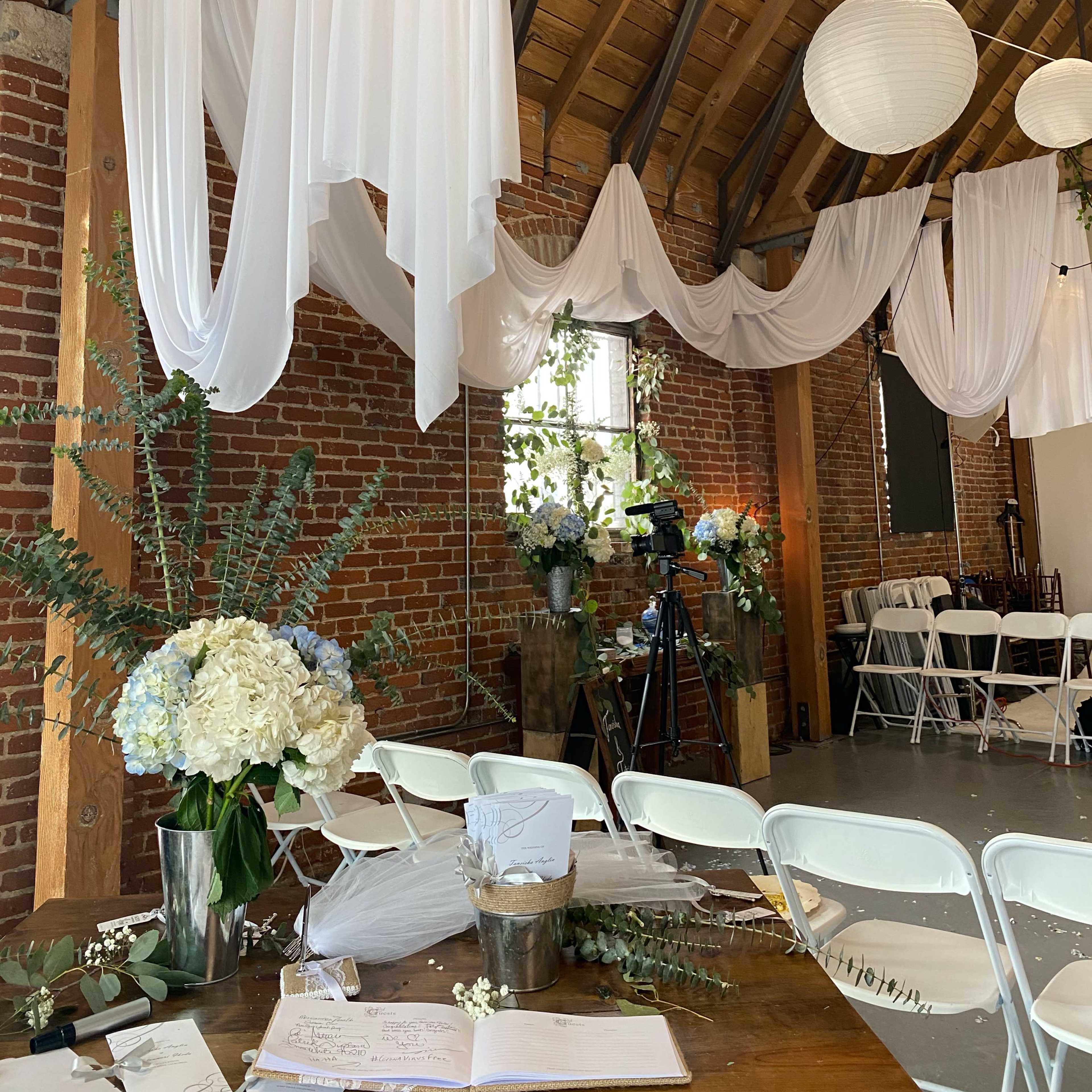 An elegantly decorated interior space with draped white fabric, floral arrangements, and chairs arranged for an event.