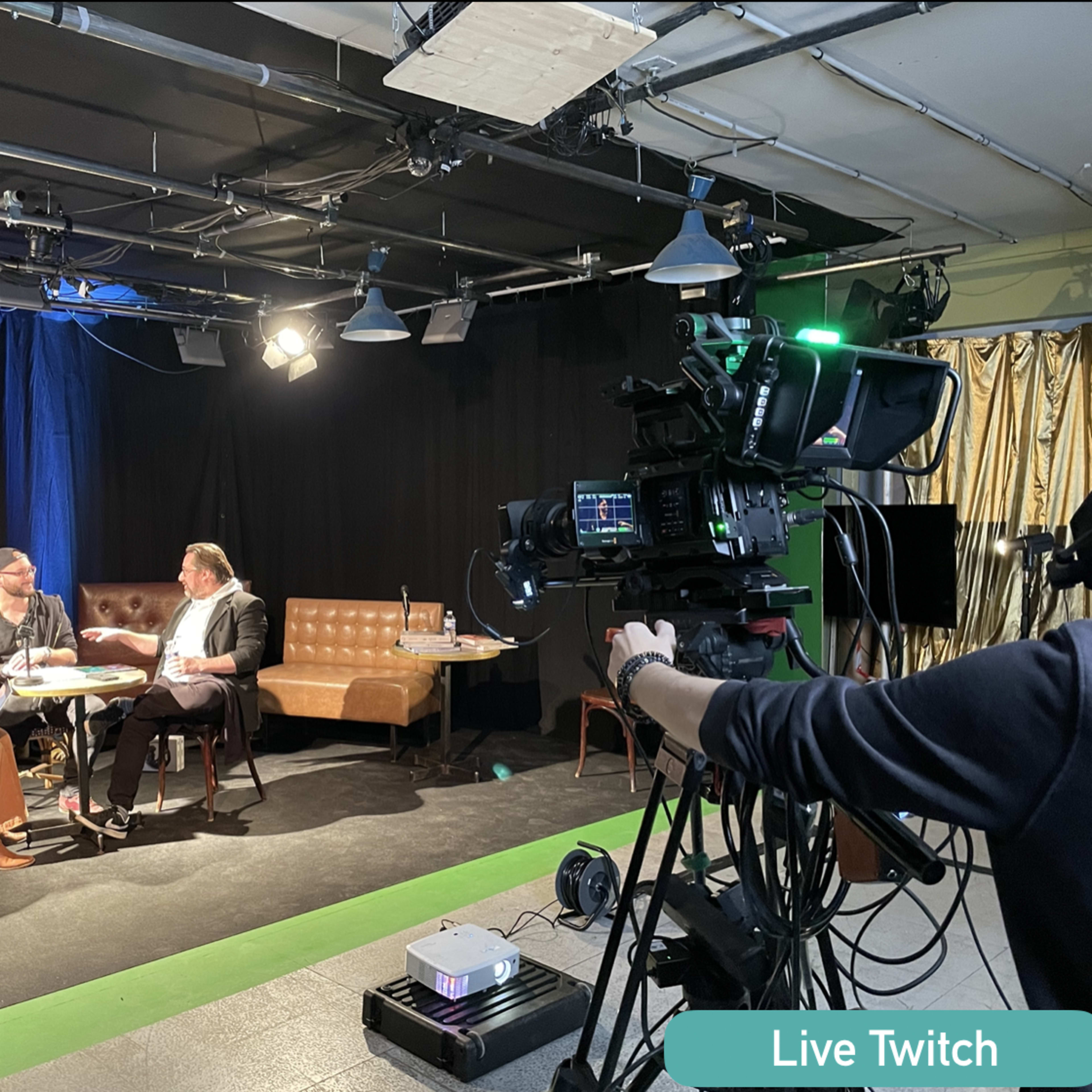 A live Twitch event is taking place on stage with a camera focused on a group of people seated and engaged in conversation.
