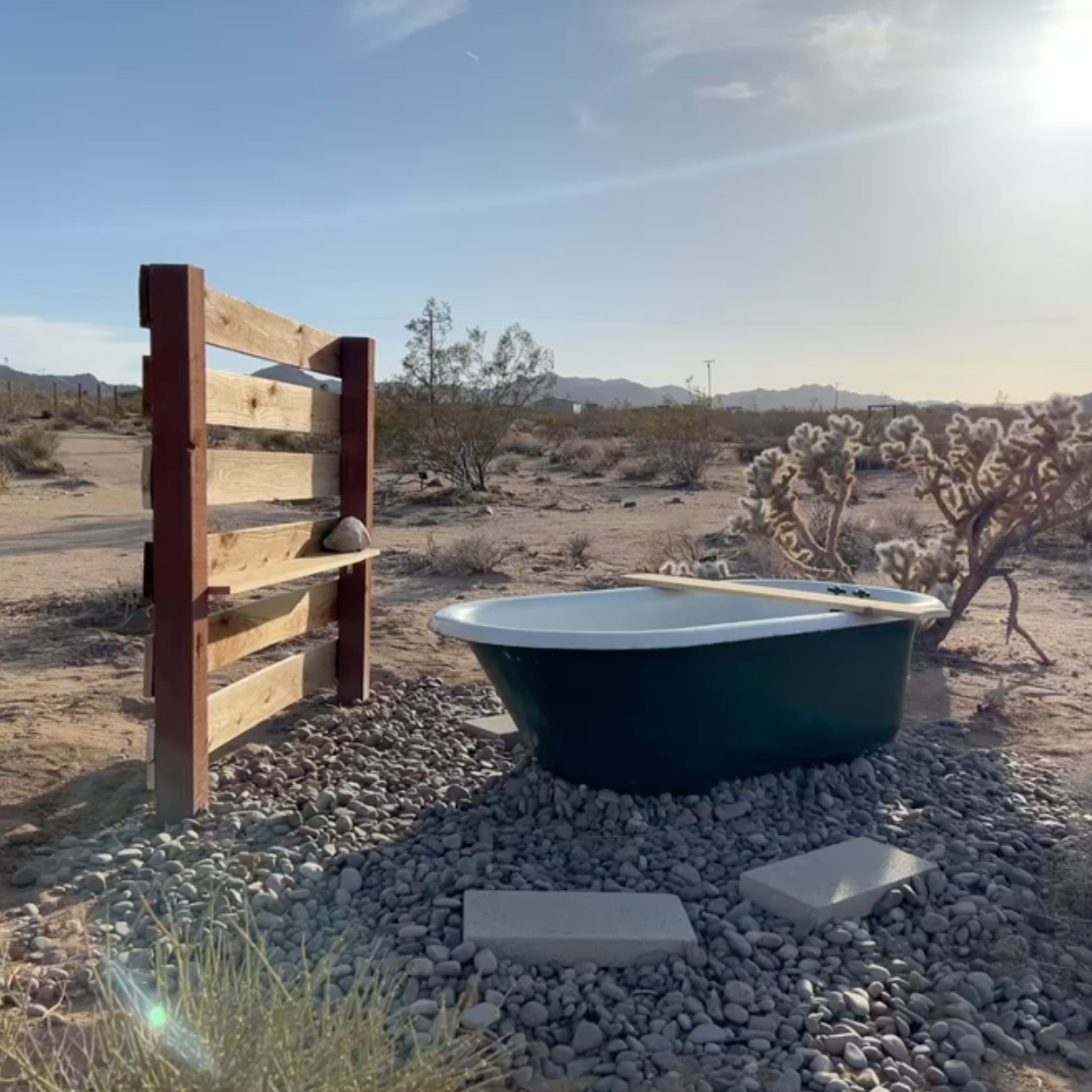 A freestanding bathtub is placed on gravel among desert vegetation with a wooden privacy screen nearby.
