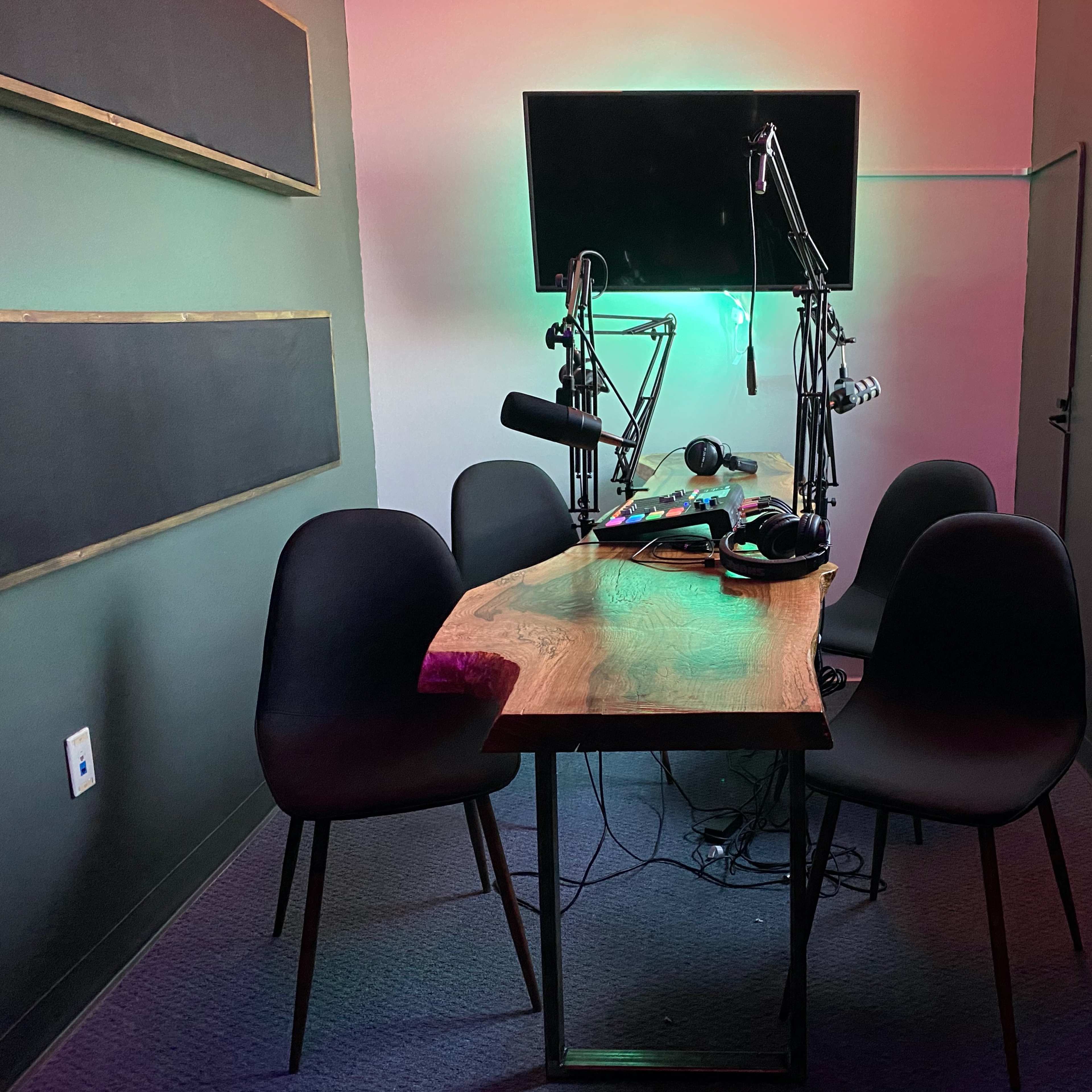 A small recording studio features a wooden table with microphones and headphones, surrounded by four black chairs and illuminated by colored lights.