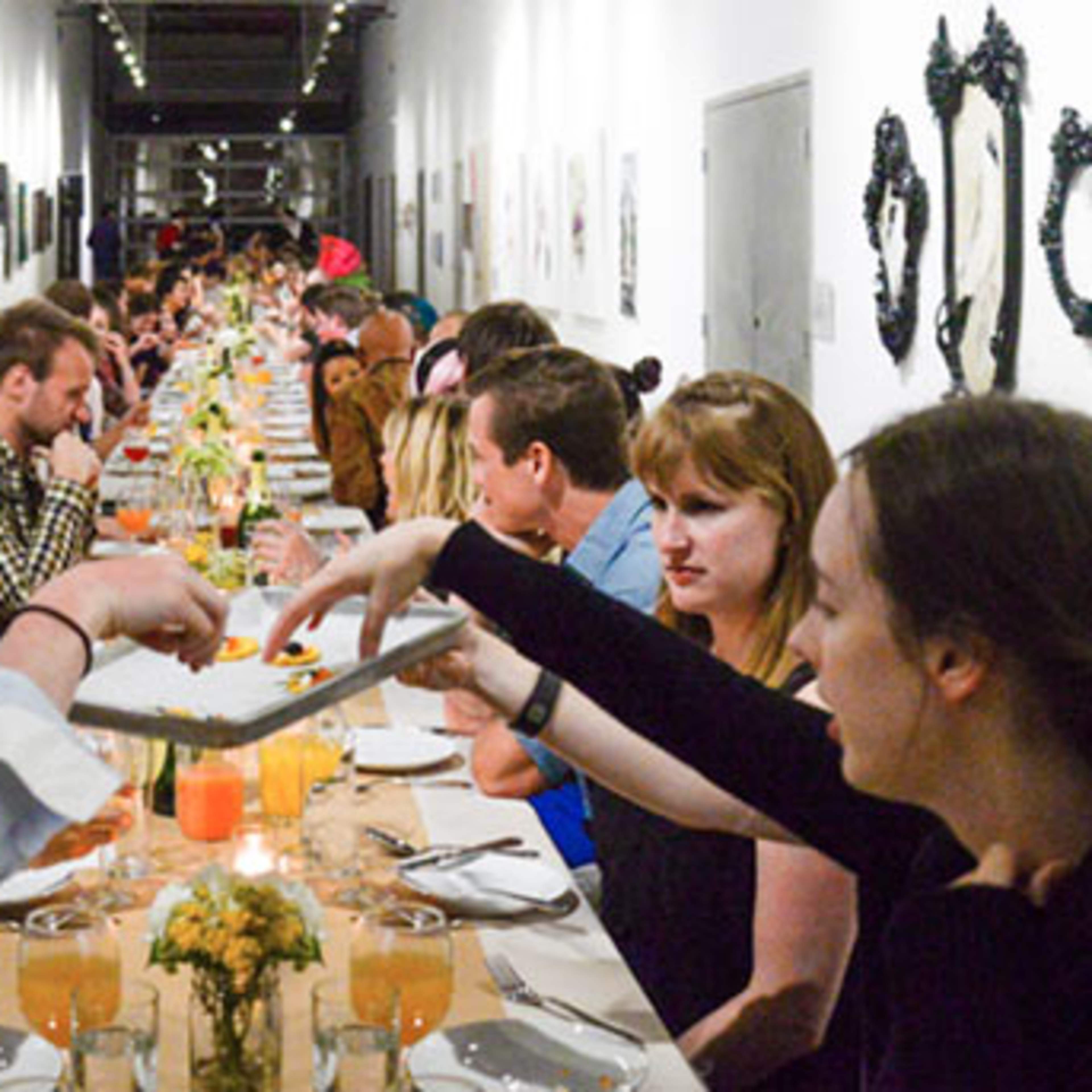 A long table filled with people dining together, surrounded by art on the walls in a well-lit gallery space.