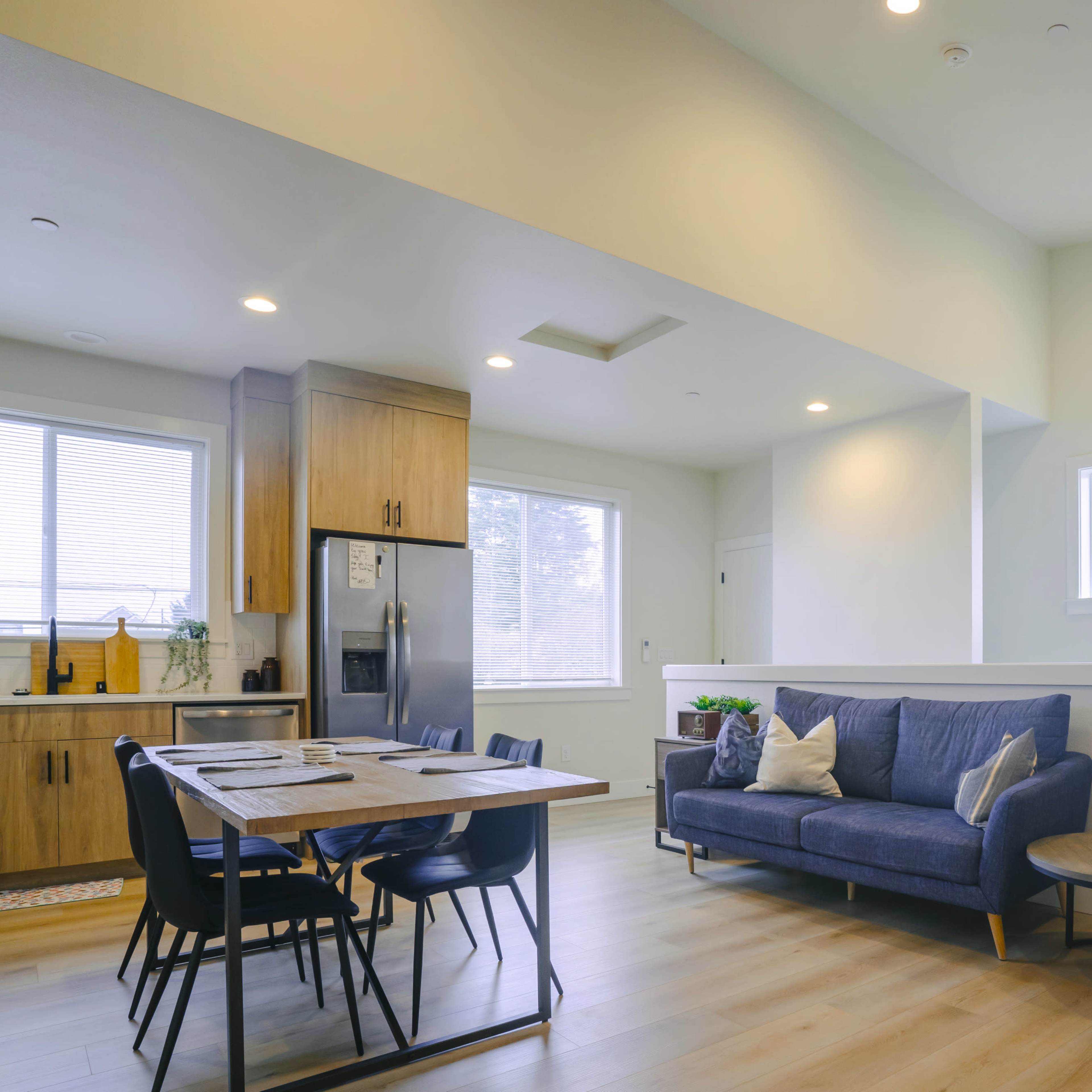 A modern, open-concept living area featuring a blue sofa, a dining table with chairs, and a kitchen with wooden cabinets.