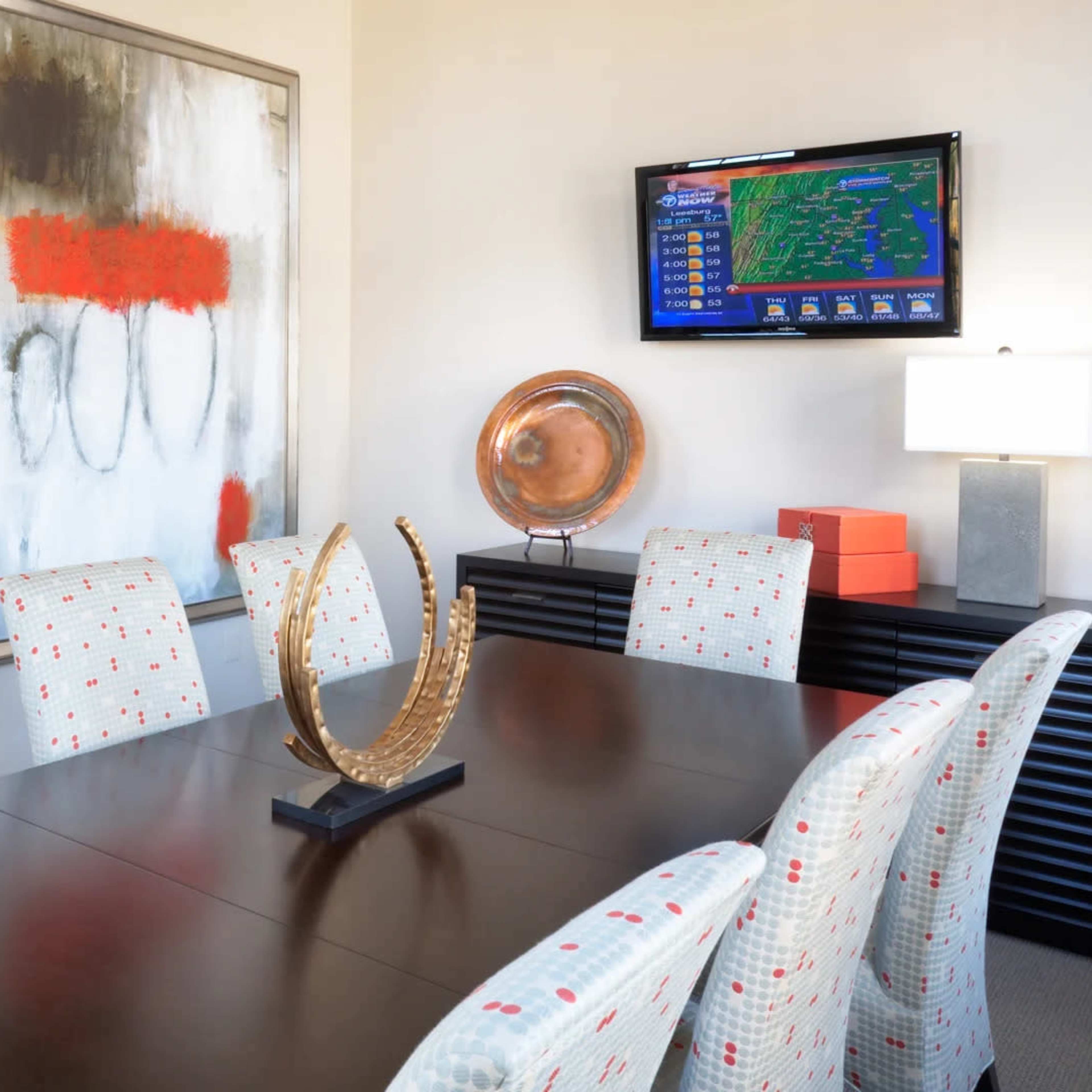 A modern conference room features a large wooden table surrounded by patterned chairs, with a colorful abstract painting on the wall and a television displaying a weather forecast.