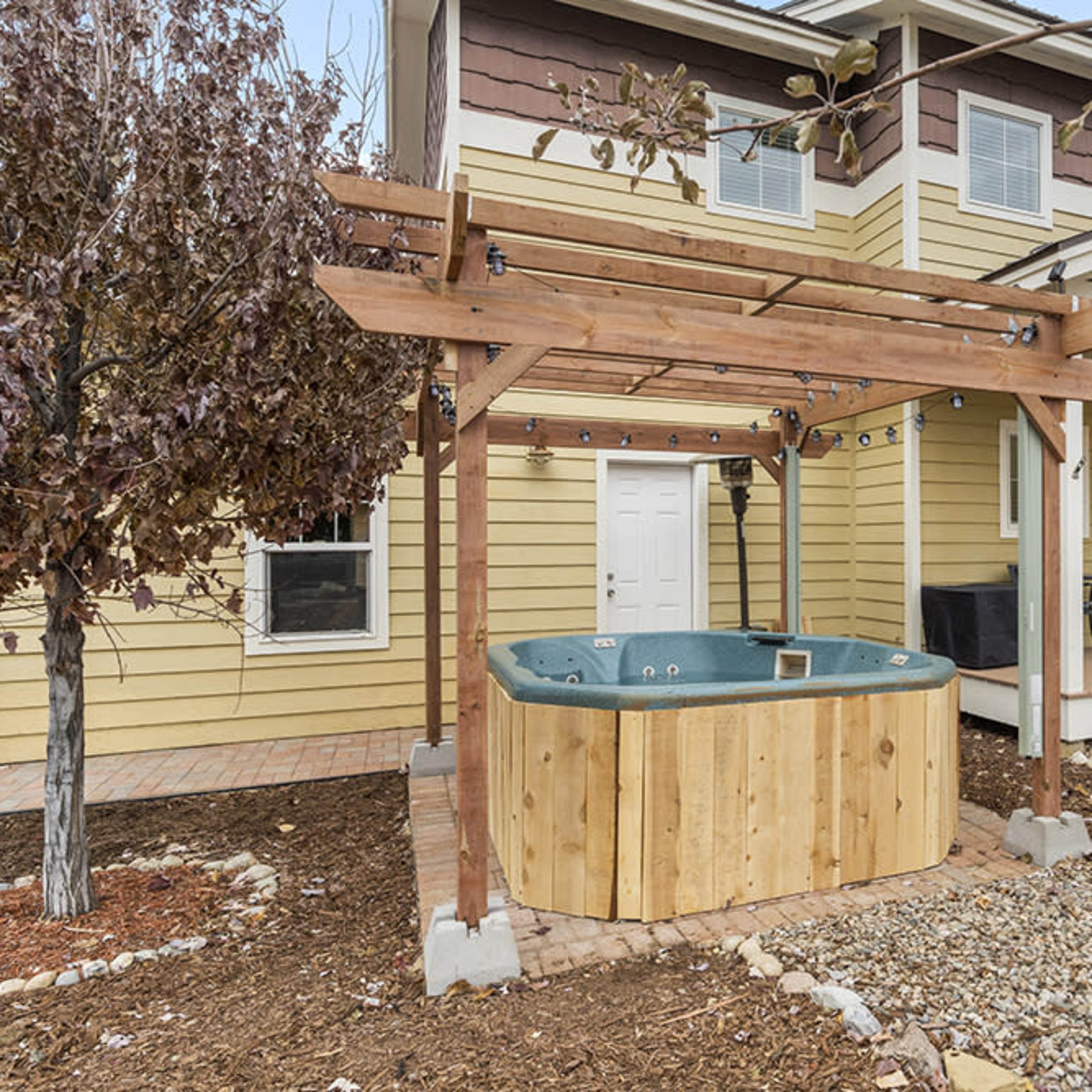 A wooden hot tub situated under a pergola in the front yard of a two-story yellow house.