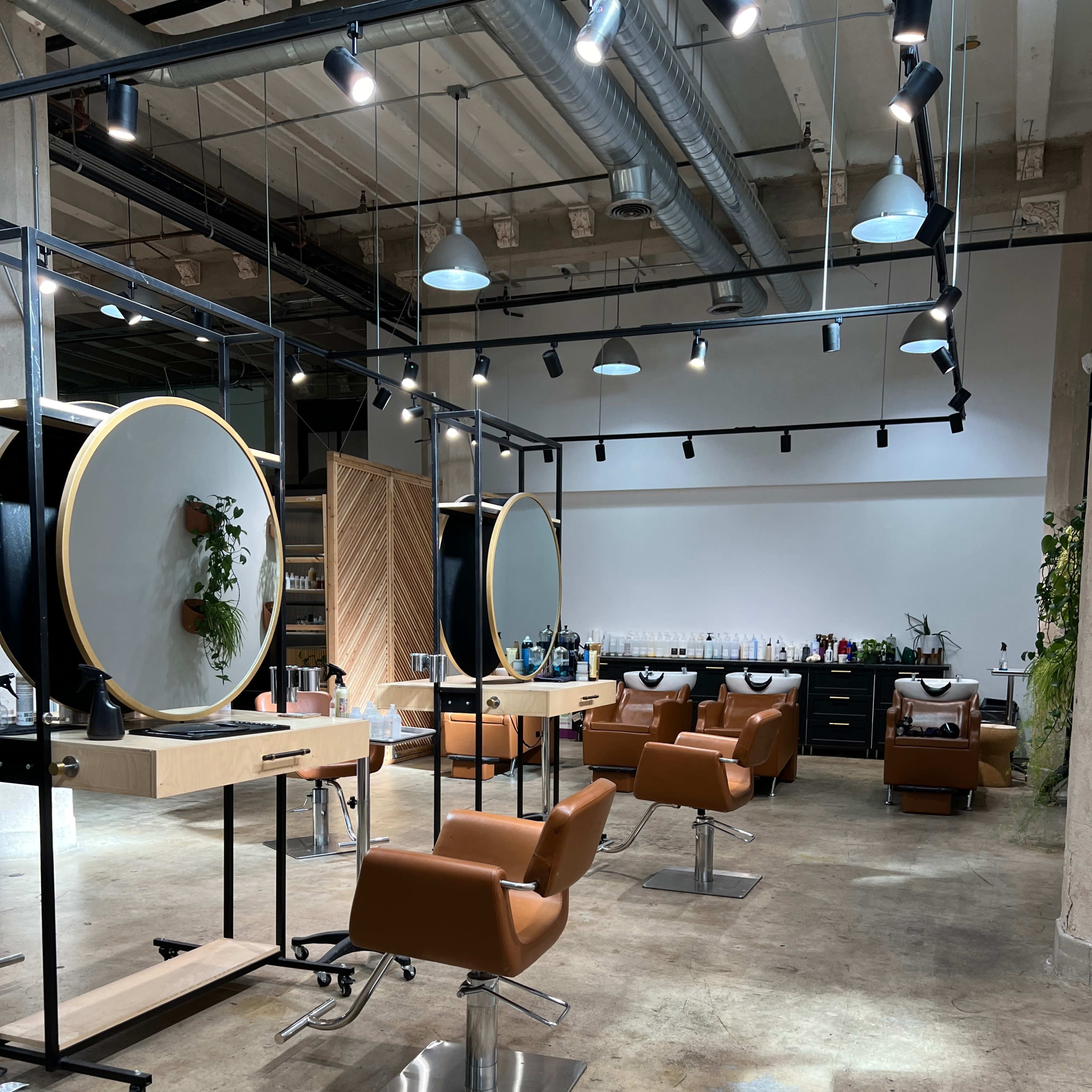 A modern salon interior with several styling stations featuring round mirrors, comfortable brown chairs, and a minimalist decor.