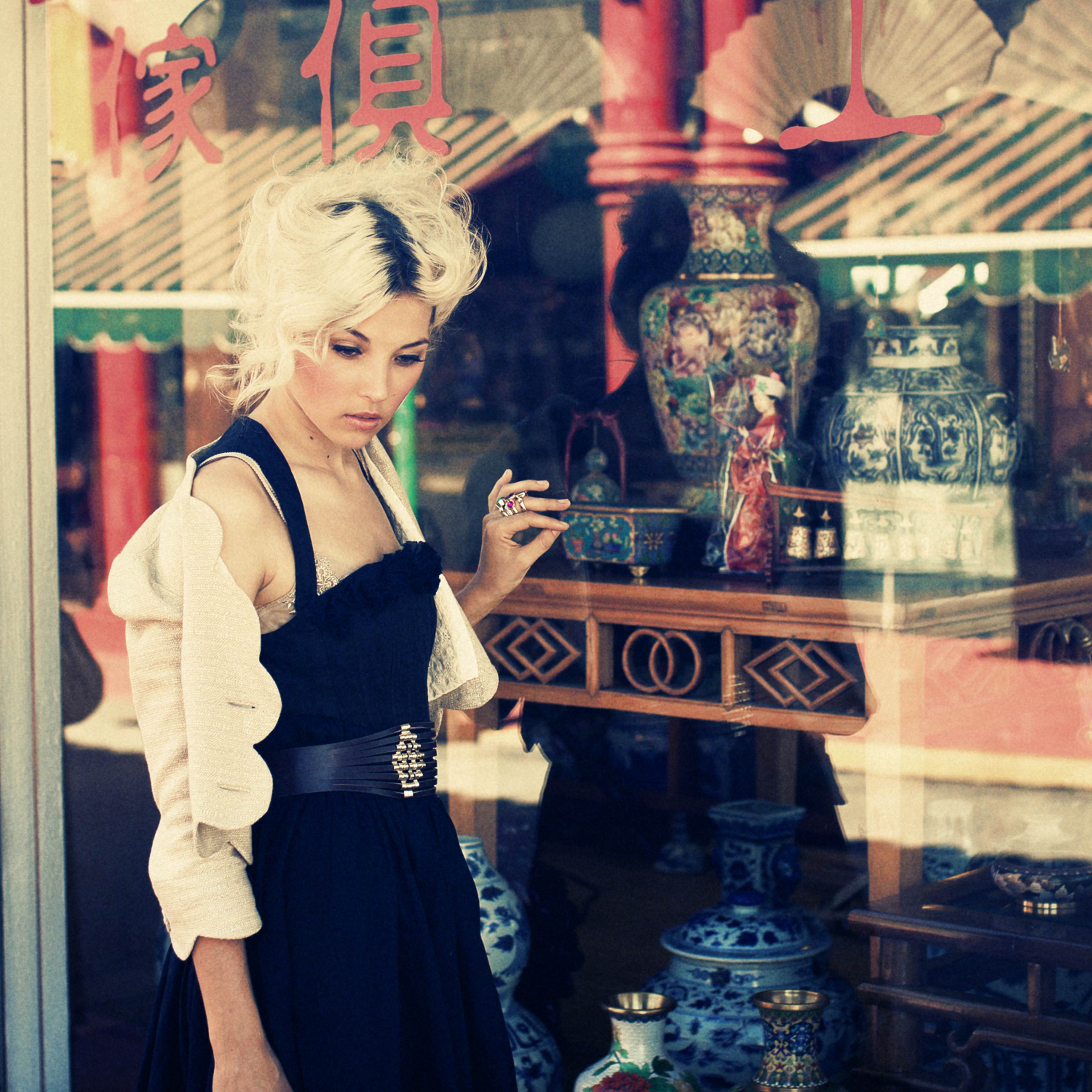 A woman in a black dress and light-colored cardigan stands near a shop window displaying intricate pottery and decorative items.