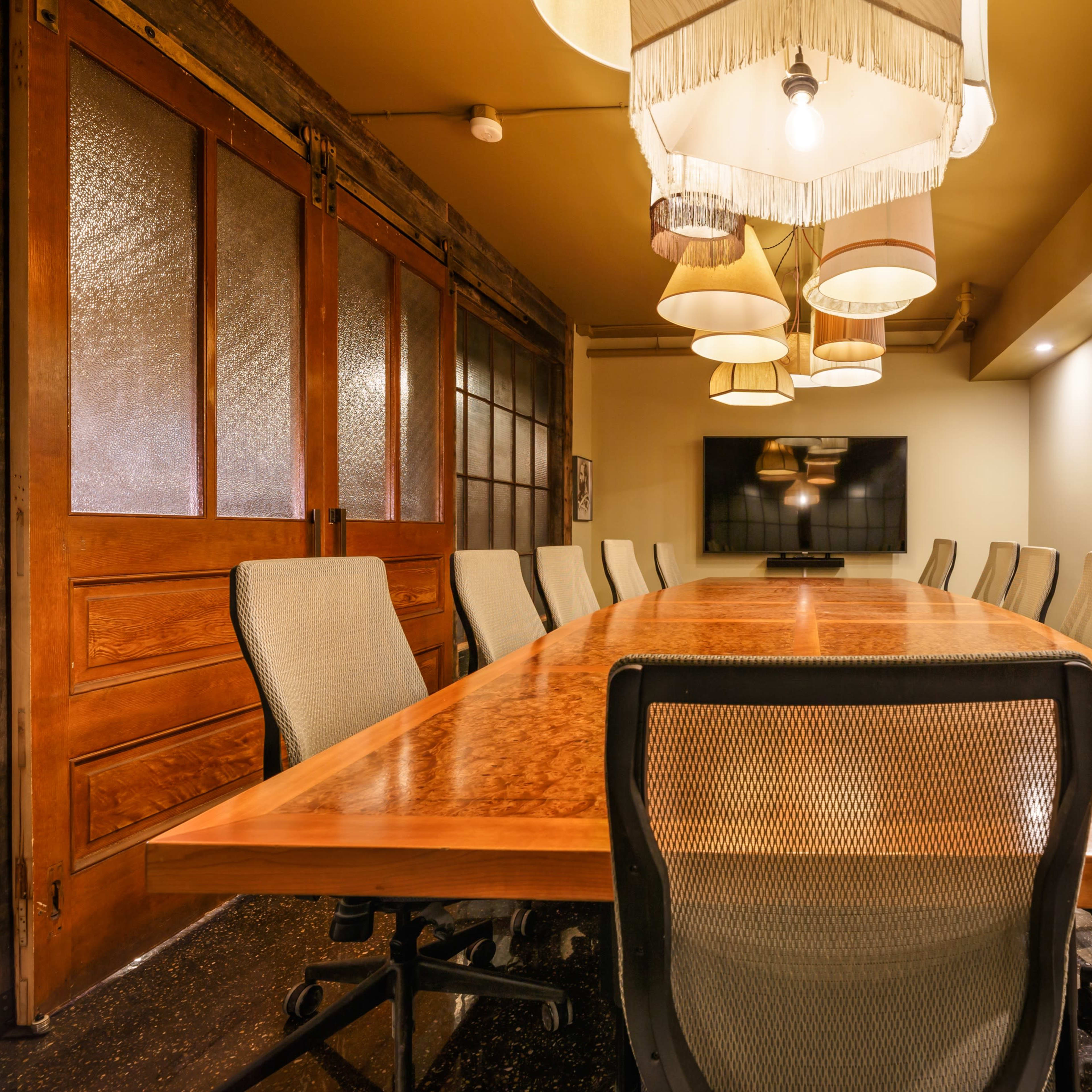 A conference room featuring a long wooden table surrounded by mesh chairs, with multiple overhead lights and a wall-mounted television.