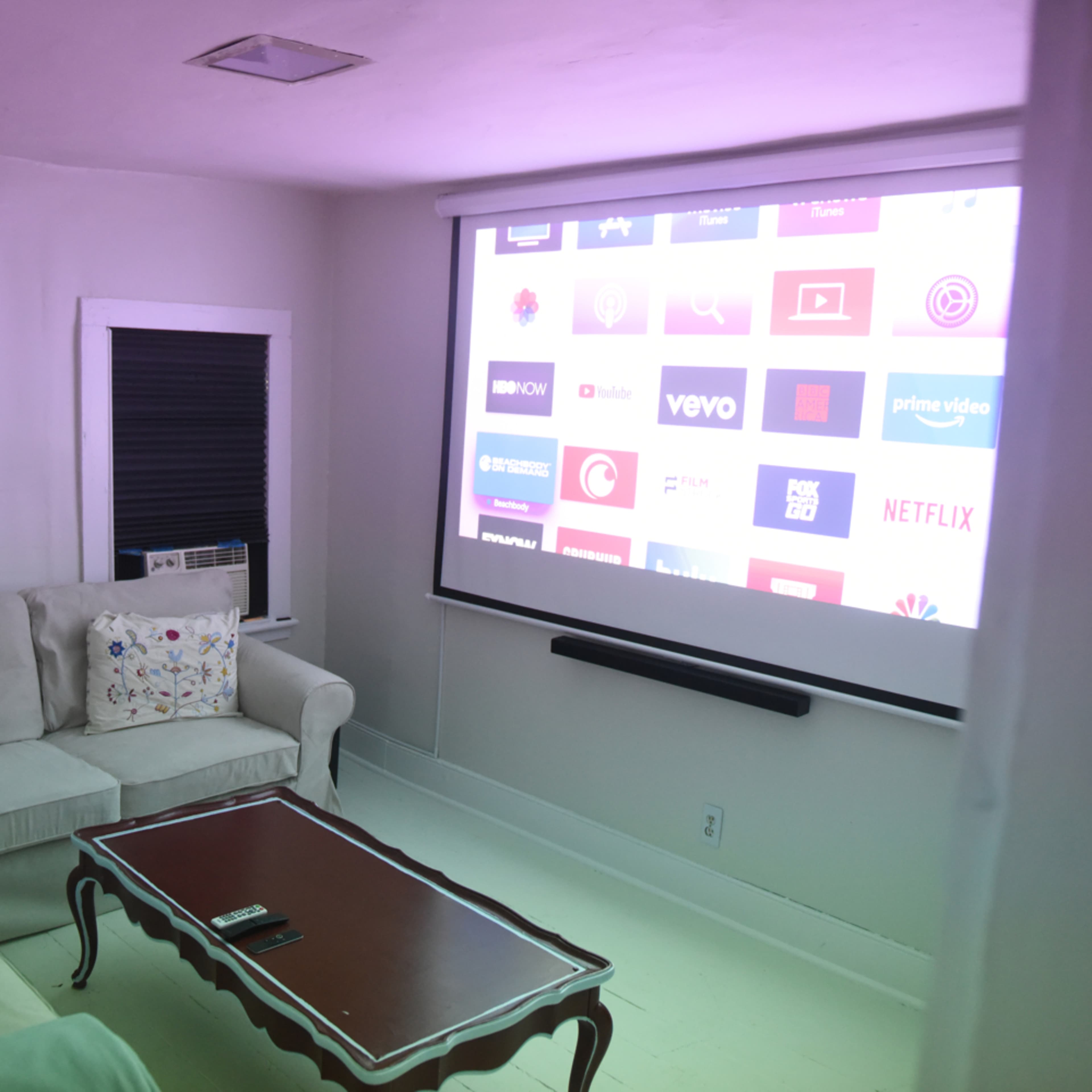 A living room features a white projector screen displaying various streaming service logos, with a beige sofa and a wooden coffee table in front.