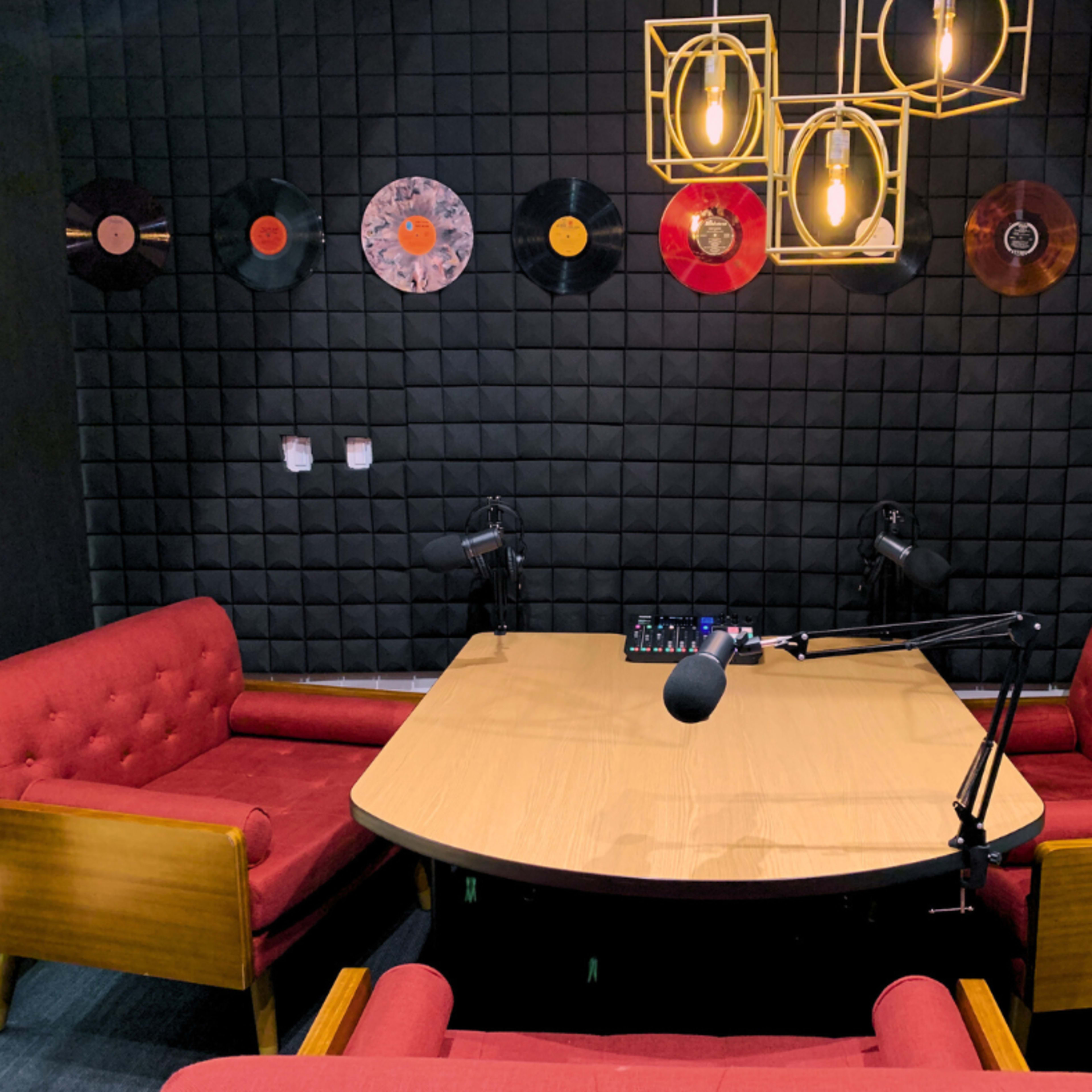 A recording studio with a round wooden table, two red upholstered chairs, and several vinyl records displayed on a black wall.