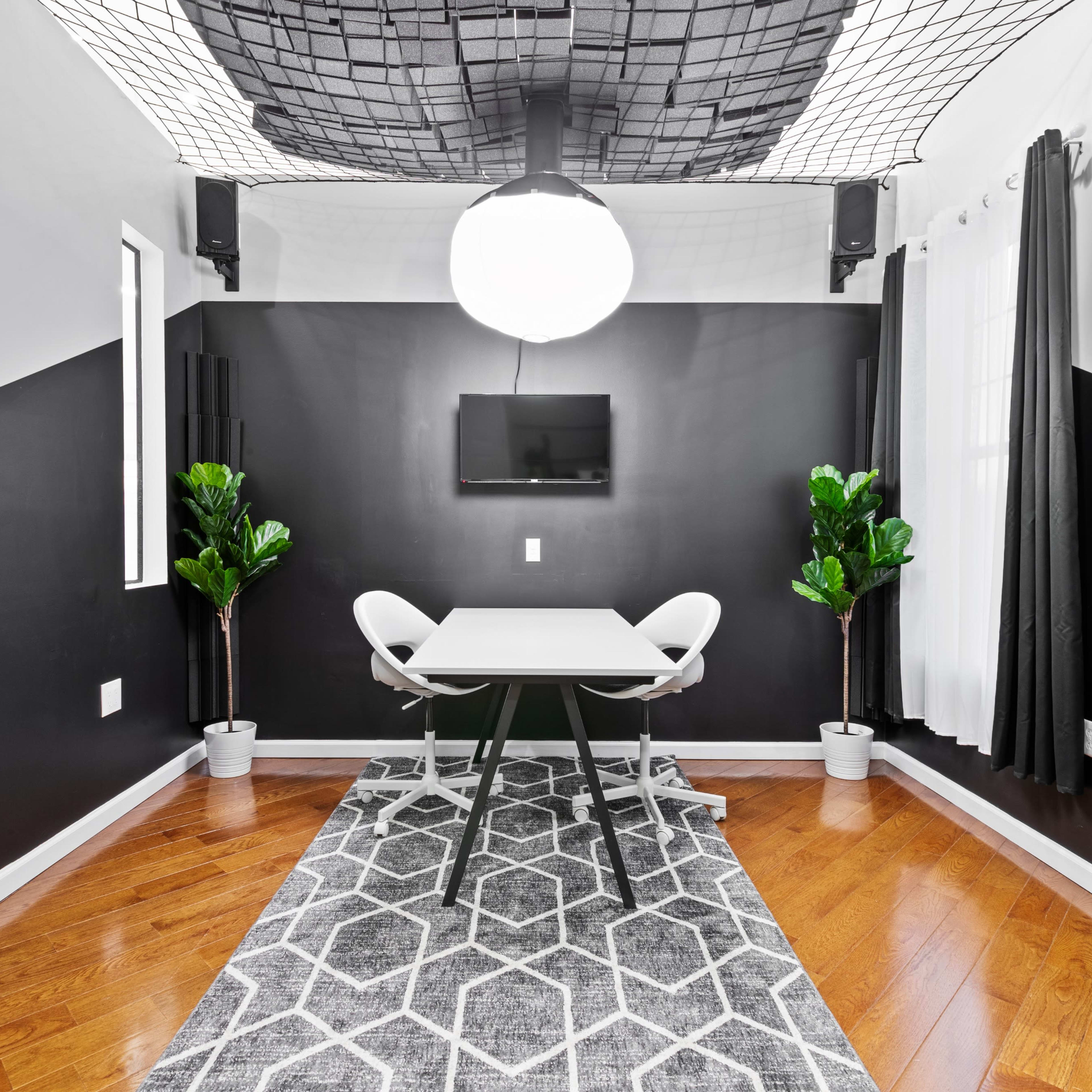 A small, modern meeting room features a table with two chairs, a large globe light fixture, and two potted plants on either side, set against black and white walls.