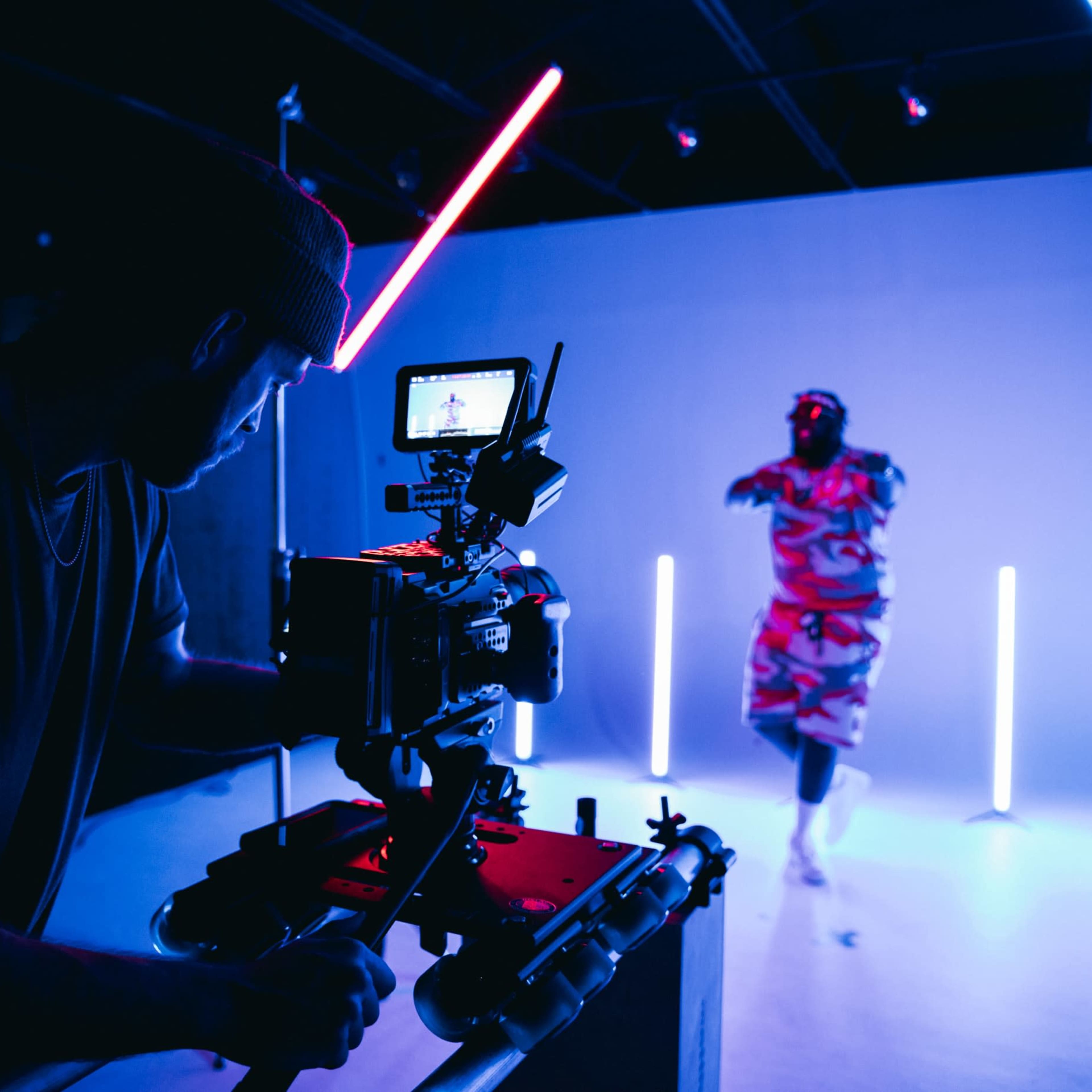 A camera operator films a dancer illuminated by neon lights in a studio setting.
