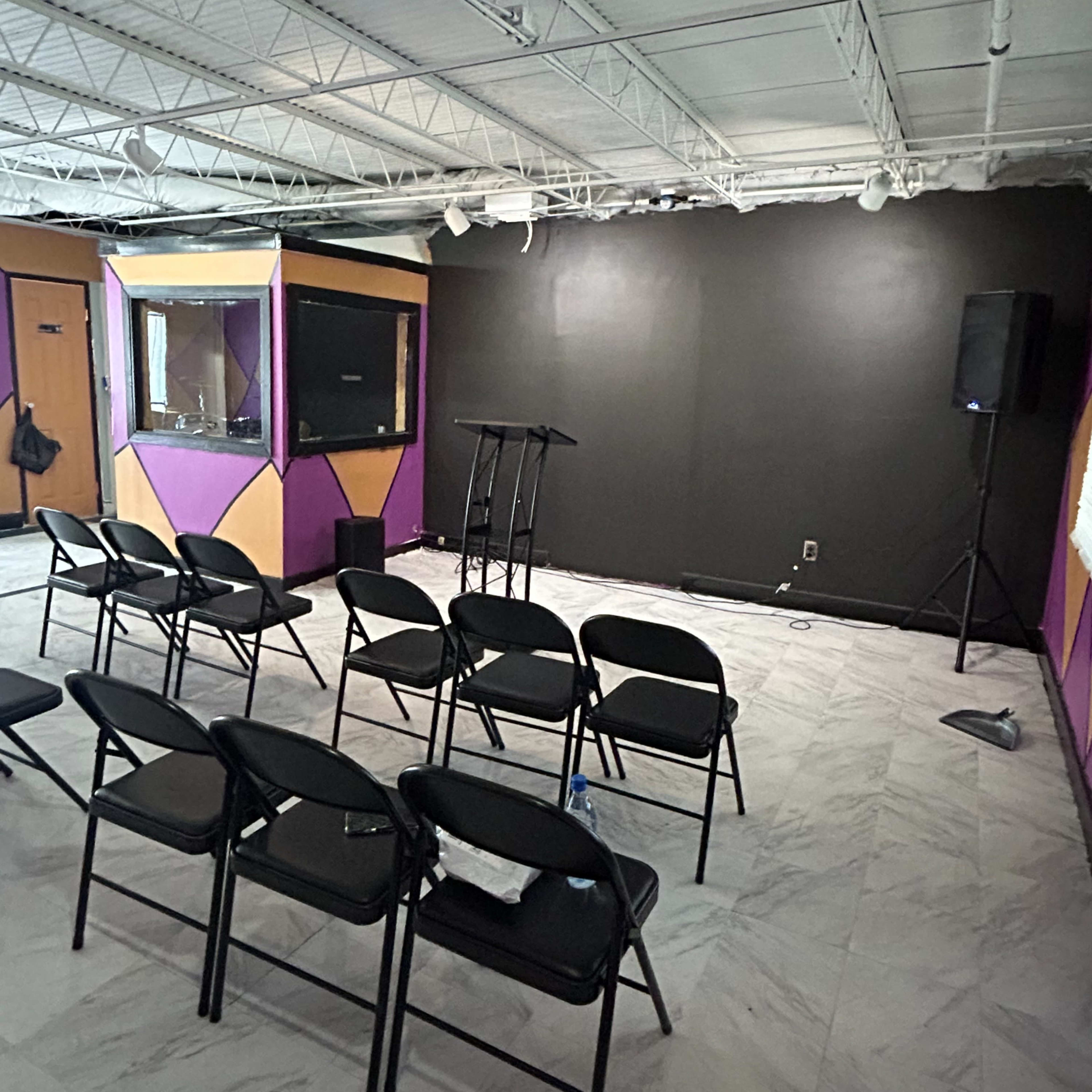 A vacant room with black walls, several rows of folding chairs, a podium, and a sound system on one side.