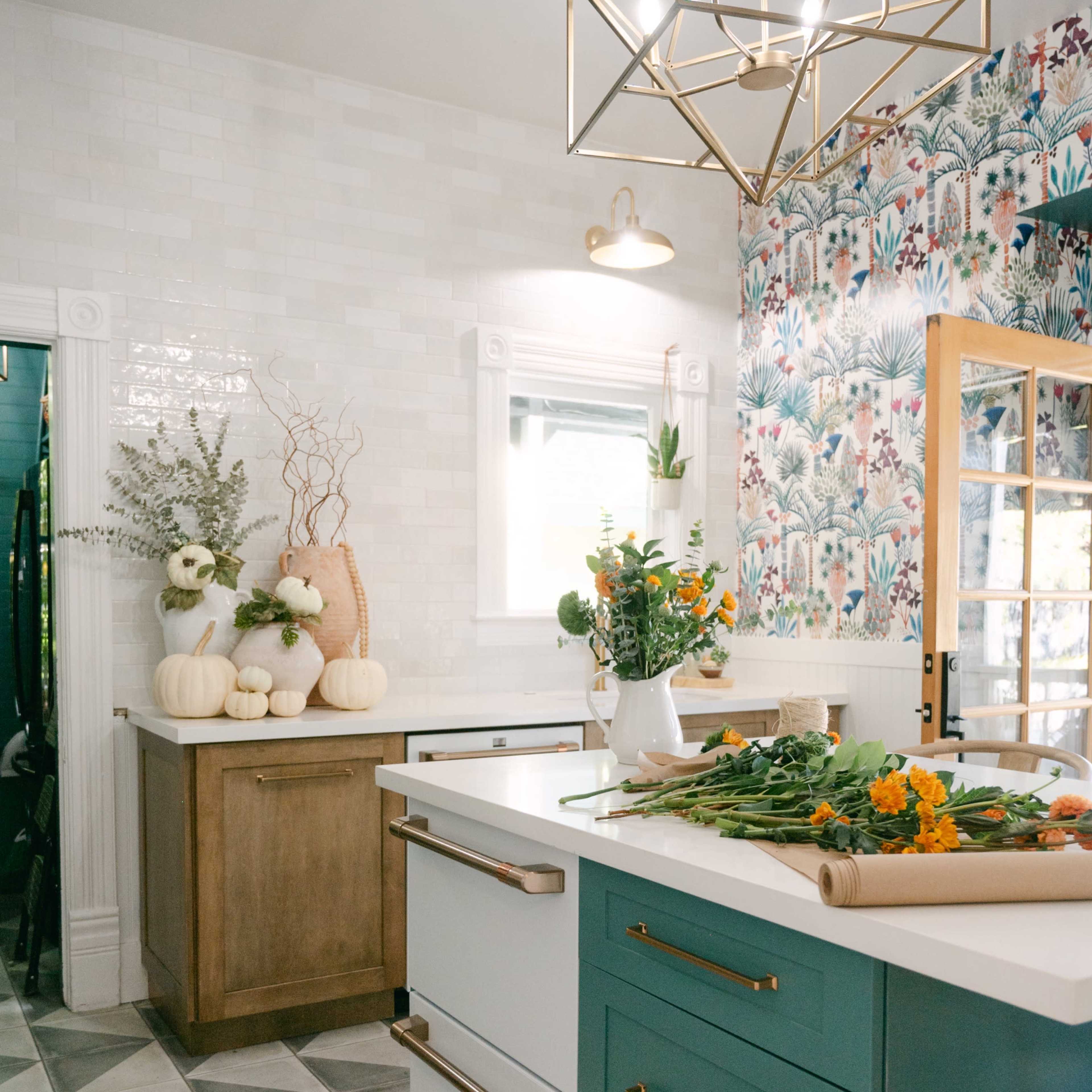 The kitchen features a spacious island with teal drawers, surrounded by a floral wallpaper and modern light fixture, alongside a wooden counter with autumn decorations.