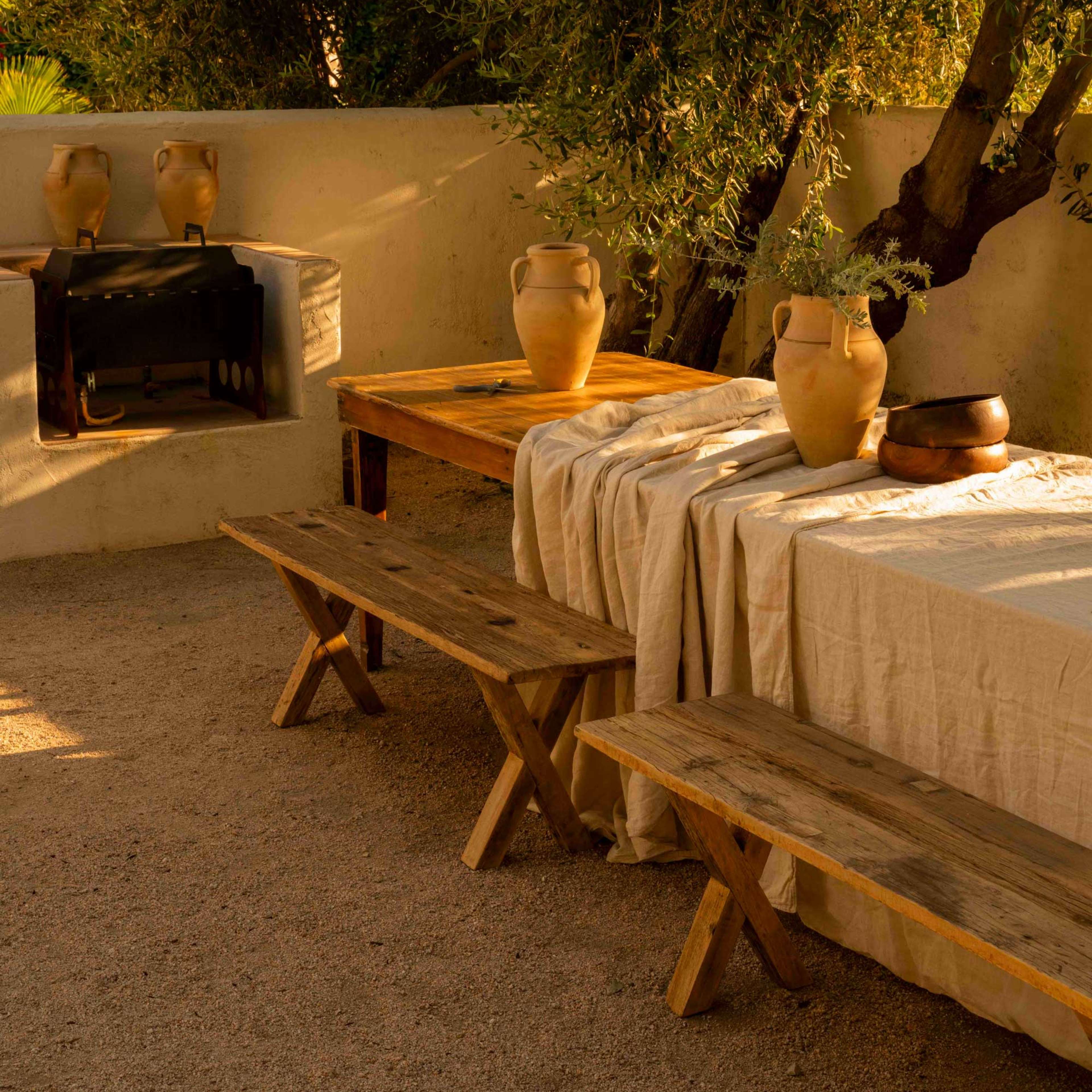 The scene features rustic wooden tables and benches under an olive tree, with clay pots arranged on the tables and a stone barbecue in the background.