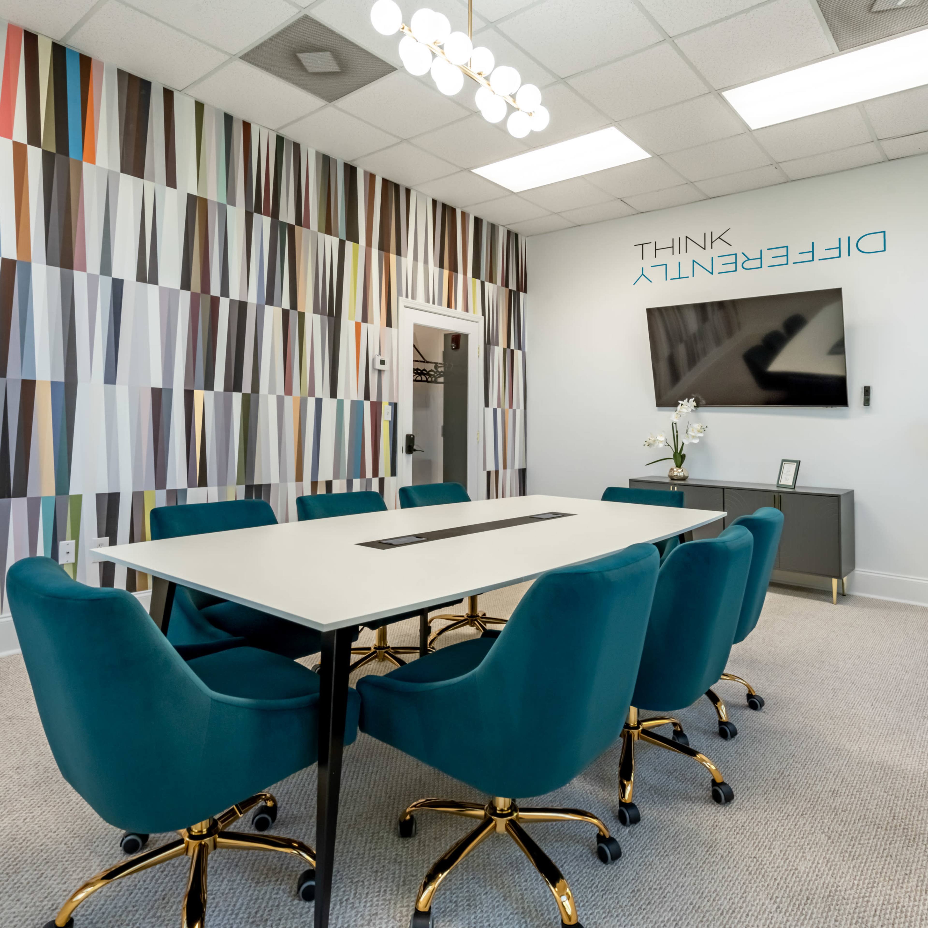 A modern conference room features a large rectangular table surrounded by teal chairs and a wall with colorful geometric artwork.